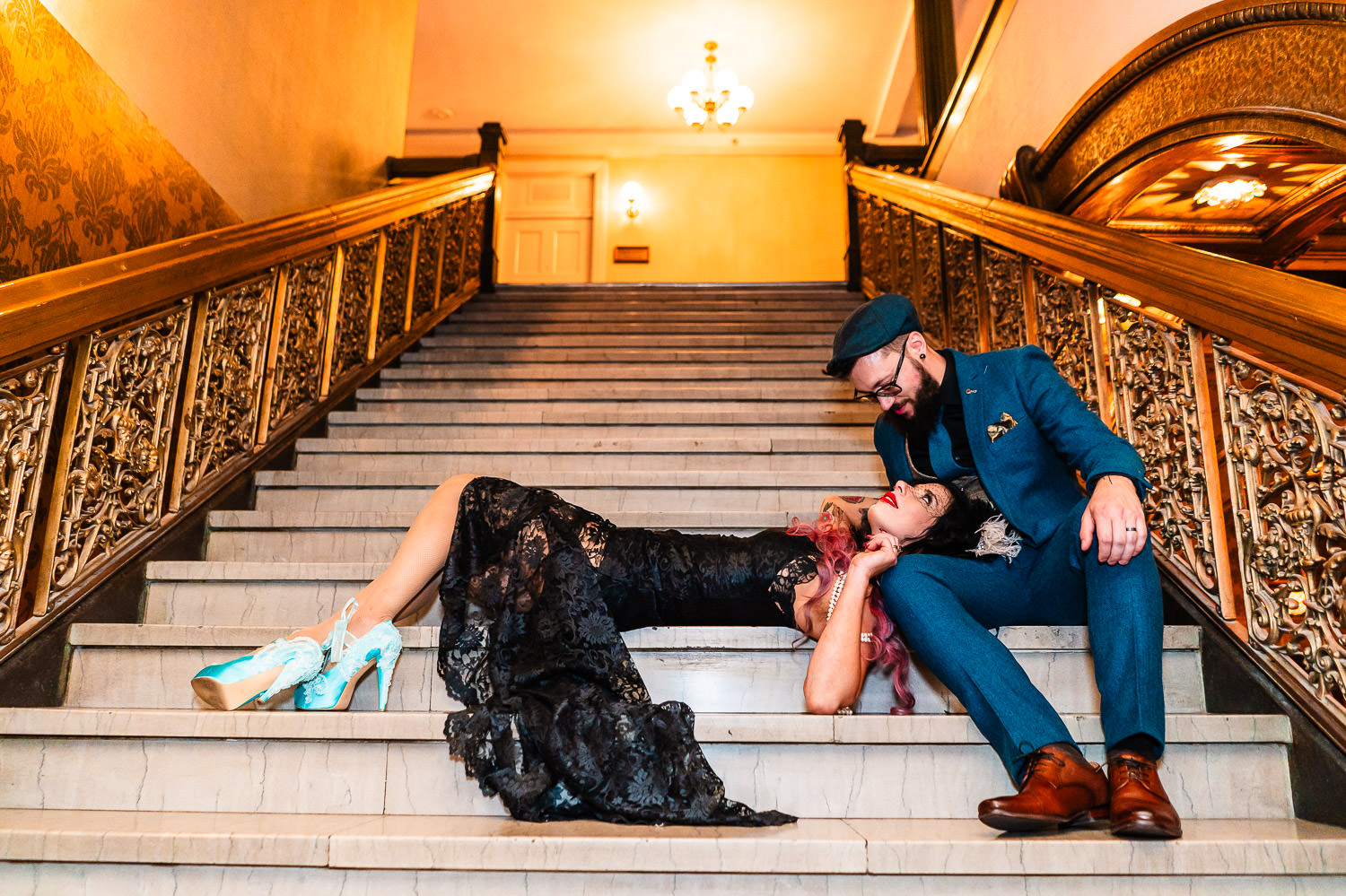 A bride in a black wedding dress lays across a step of an ornate staircase and into the lap of her groom as they gaze in each other's eyes during their 1920s-themed Brown Palace wedding in Denver, Colorado.