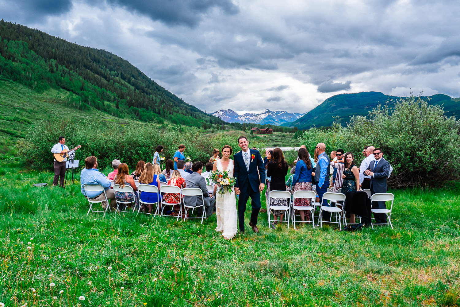 A newlywed couple walking down the aisle outdoors with guests on either side and a mountainous backdrop.