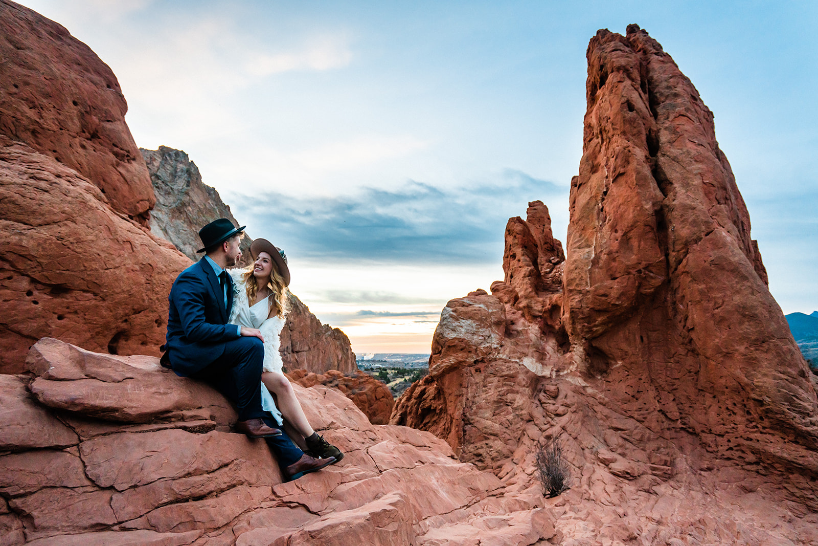 A couple in stylish attire smiles at one another and sits on a rocky outcrop near the landmark Sleeping Giant in Garden of the Gods during their Colorado Springs adventure photography session.