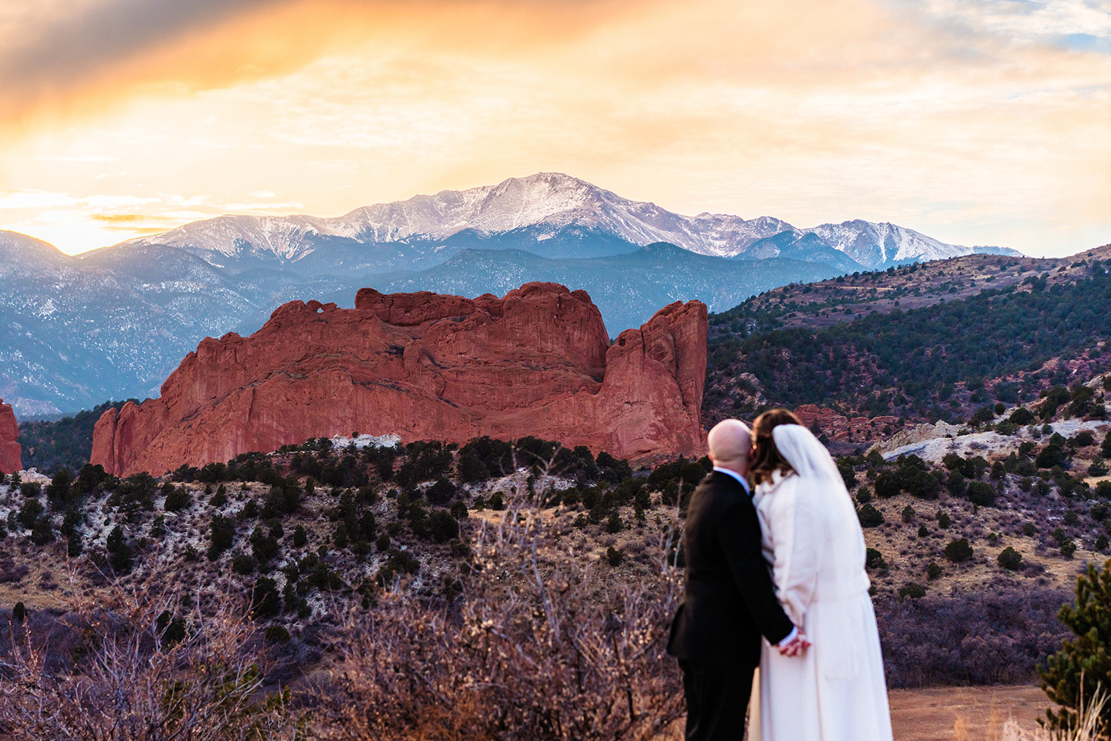 A bride and groom kiss with a backdrop of red rock formations and snow-capped mountains at sunset during their Garden of the Gods elopement.