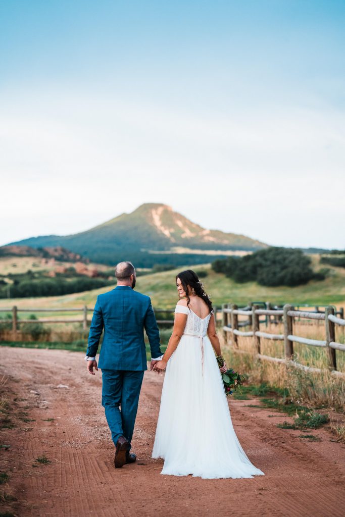 newlyweds hold hands and walk down a dirt road toward the mountains