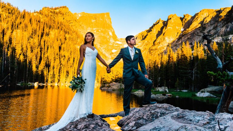 Husband and wife stand hand in hand in front of a mountain lake during sunrise