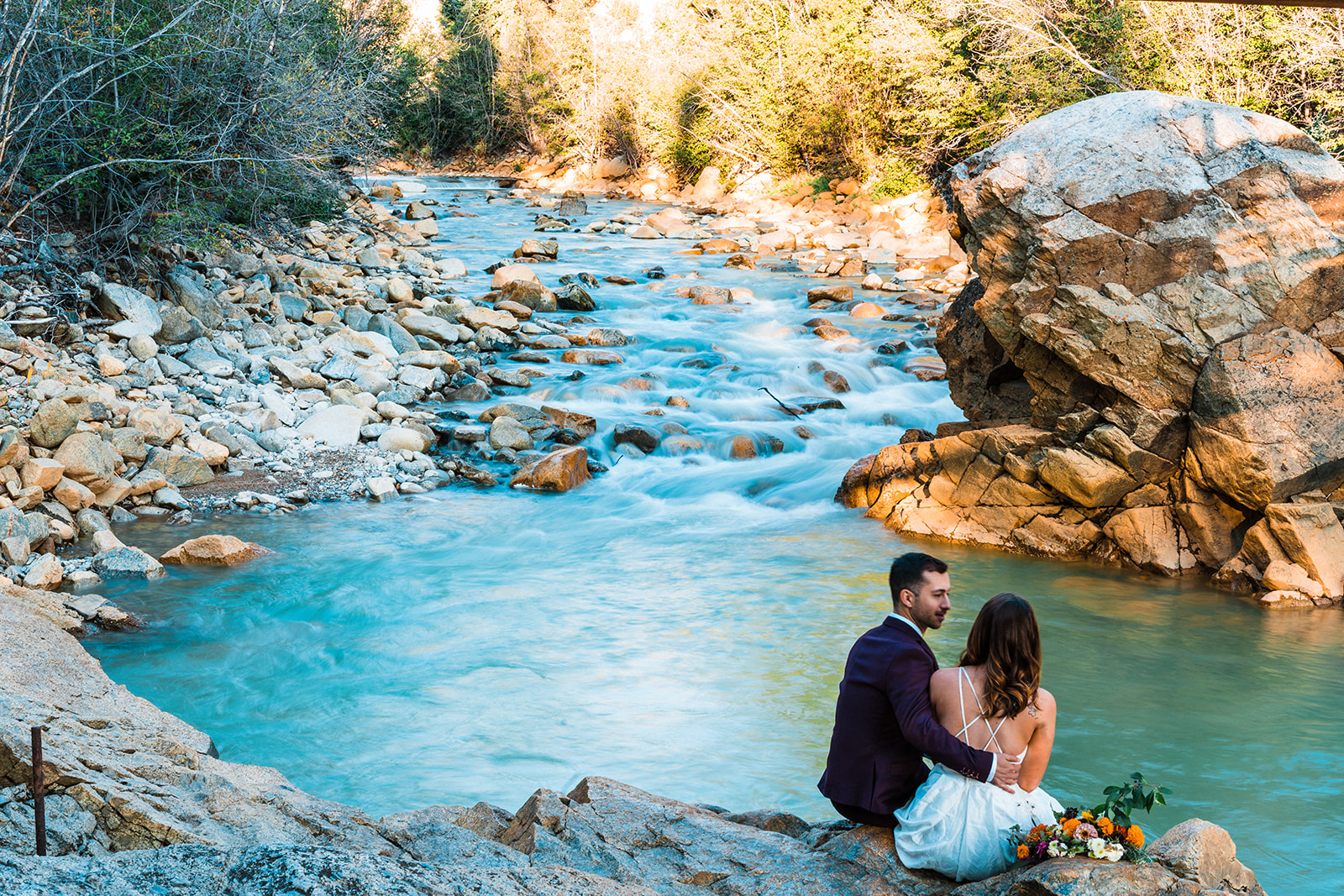 Newlyweds sit by a turquoise river with gray stones about, the man in a suit and the woman in a  white dress during their Buena Vista elopement in Colorado. 