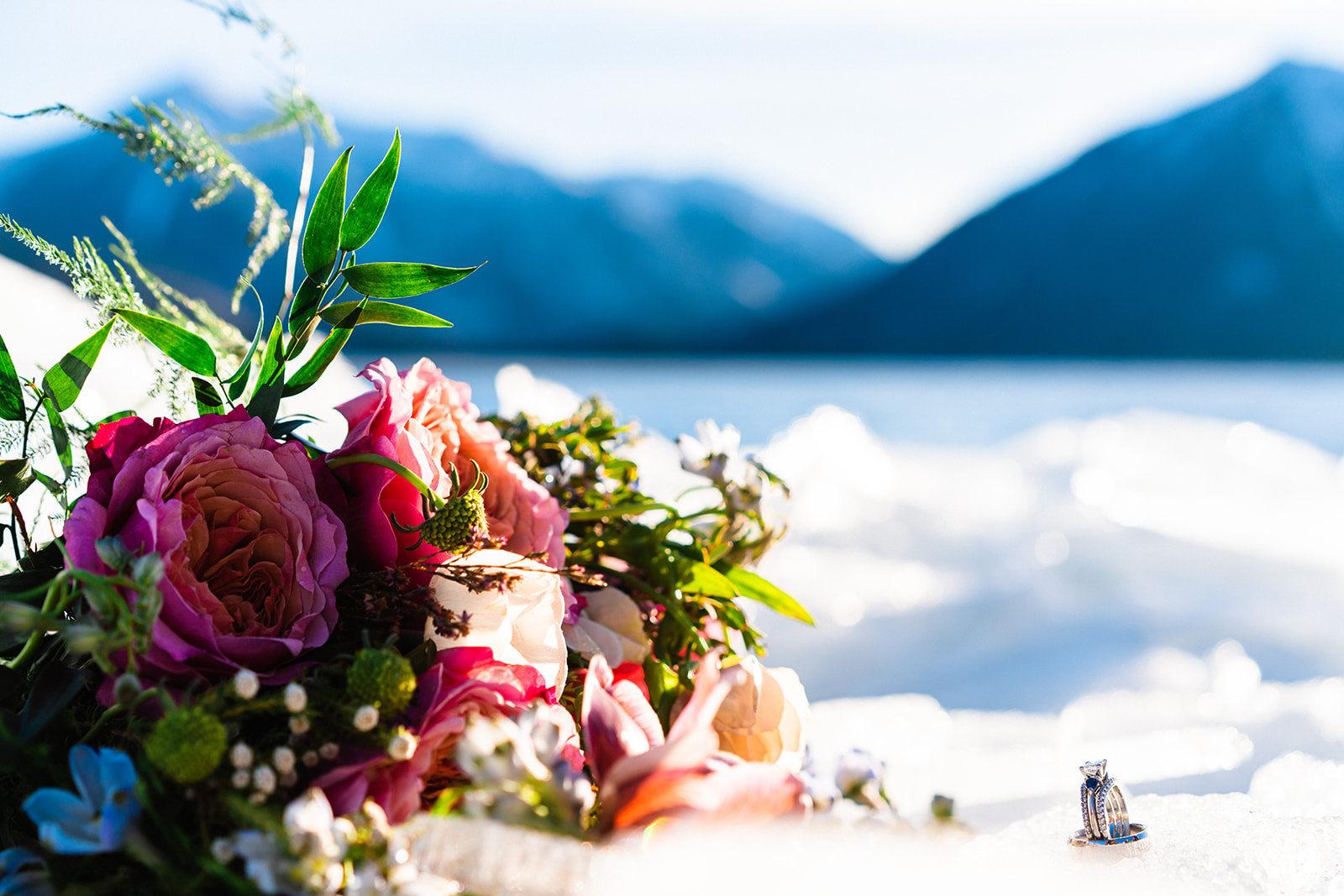 A colorful bouquet and wedding rings sit beside an icy mountain lake under a bright blue sky during a Buena Vista elopement. 