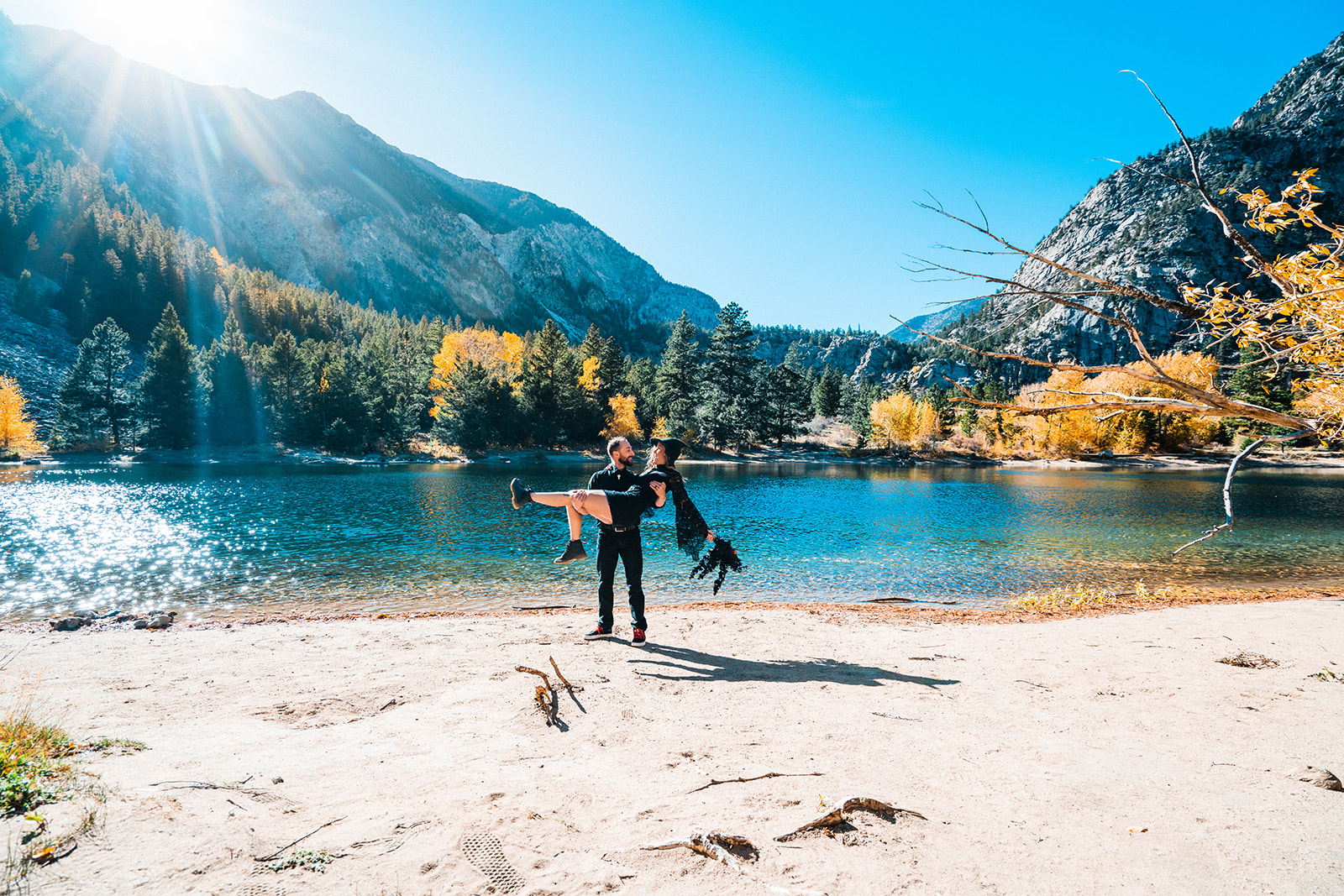 A groom in black attire holding his bride in a black elopement dress on a sandy lakeshore, 
surrounded by autumn trees and mountains under a clear blue sky during their Buena Vista elopement.