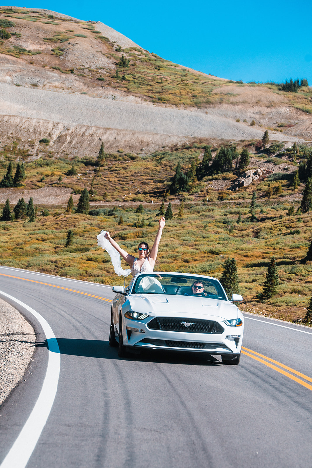A joyful newlywed couple in a white Mustang convertible car driving along Cottonwood Pass with the bride raising her arms exuberantly during their Buena Vista elopement in Colorado.