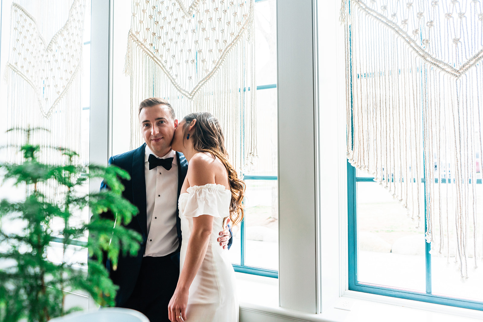 A bride kissing a groom on the cheek by a window adorned with macramé hangings during their Buena Vista elopement in Colorado at the Surf Hotel.
