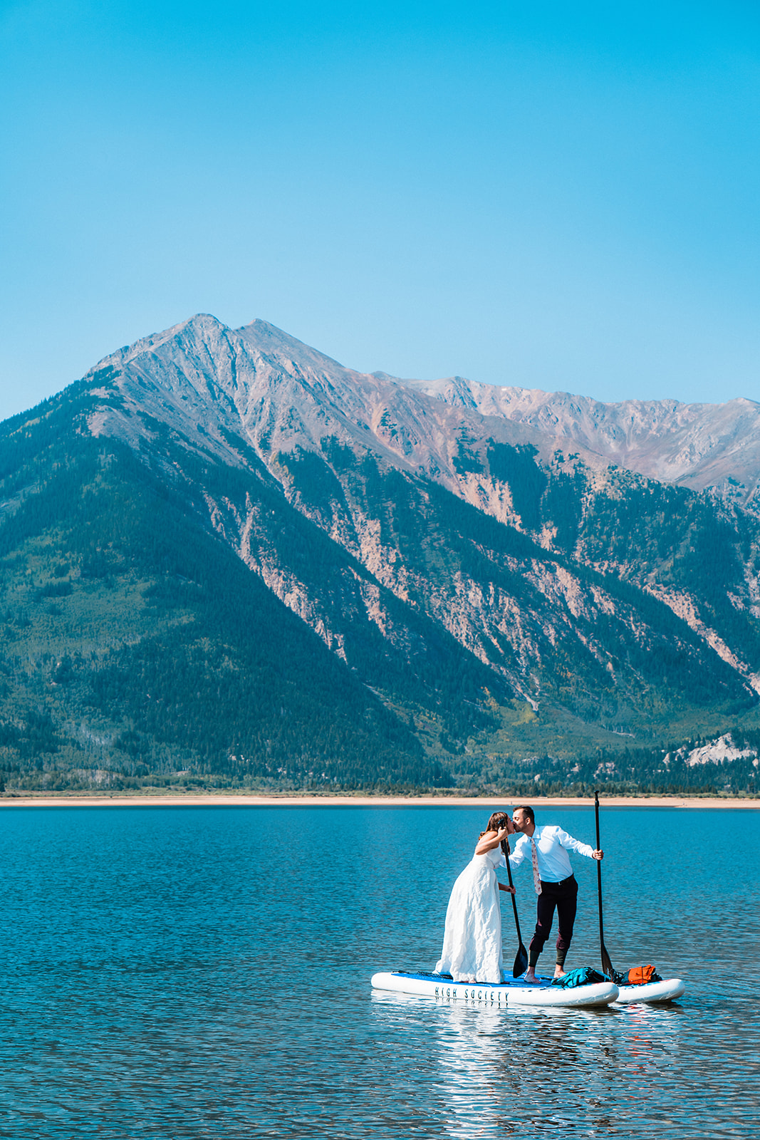 A bride in a white dress and a groom in a shirt and tie stand on separate paddle boards and kiss on a calm lake with mountains in the background during their Buena Vista elopement at Twin Lakes in Colorado.