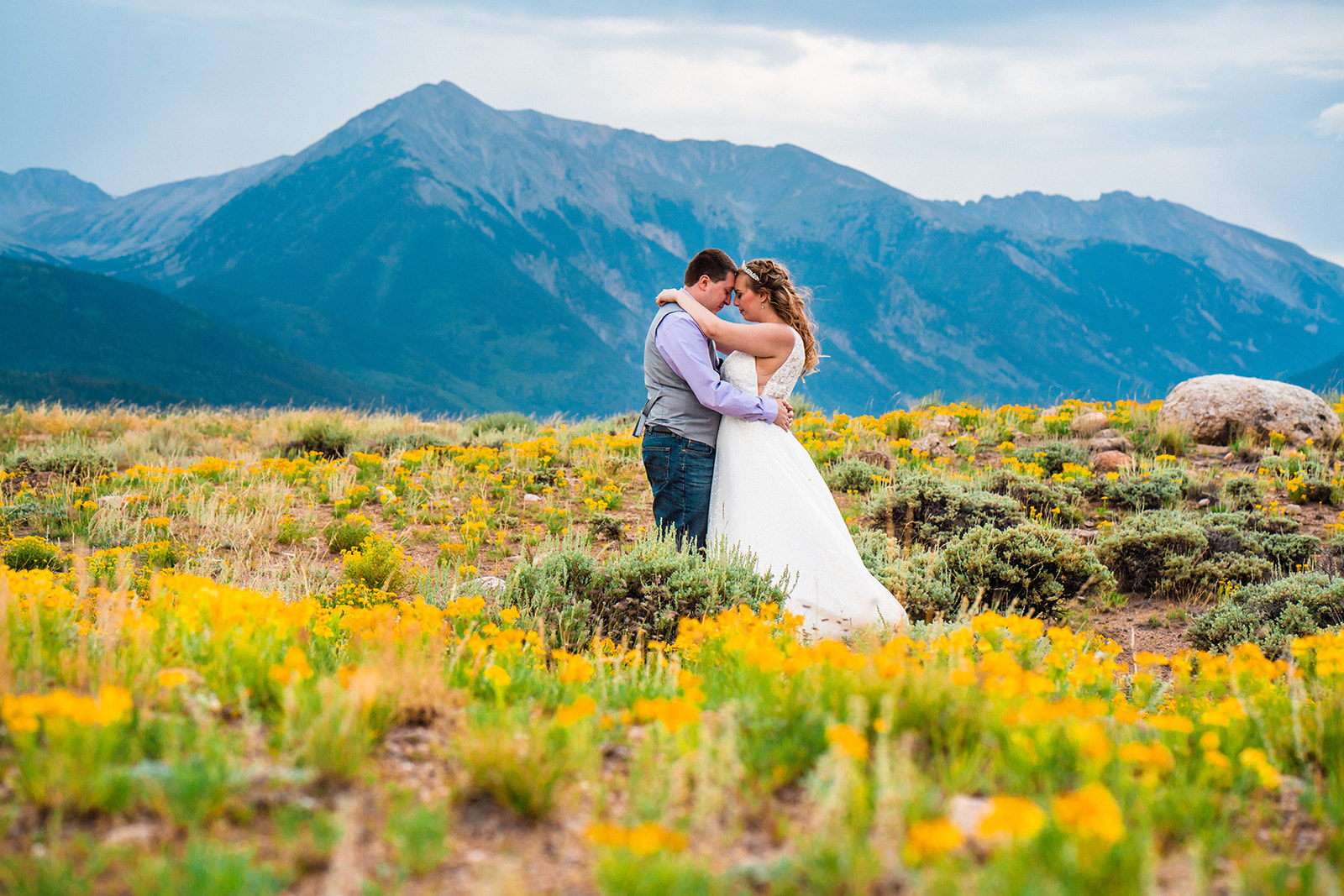 A bride and groom snuggle in mountain pasture adorned with hundreds of yellow wildflowers with tall and wide mountains behind them under a cloudy sky at Twin Lakes during their Buena Vista elopement in Colorado.