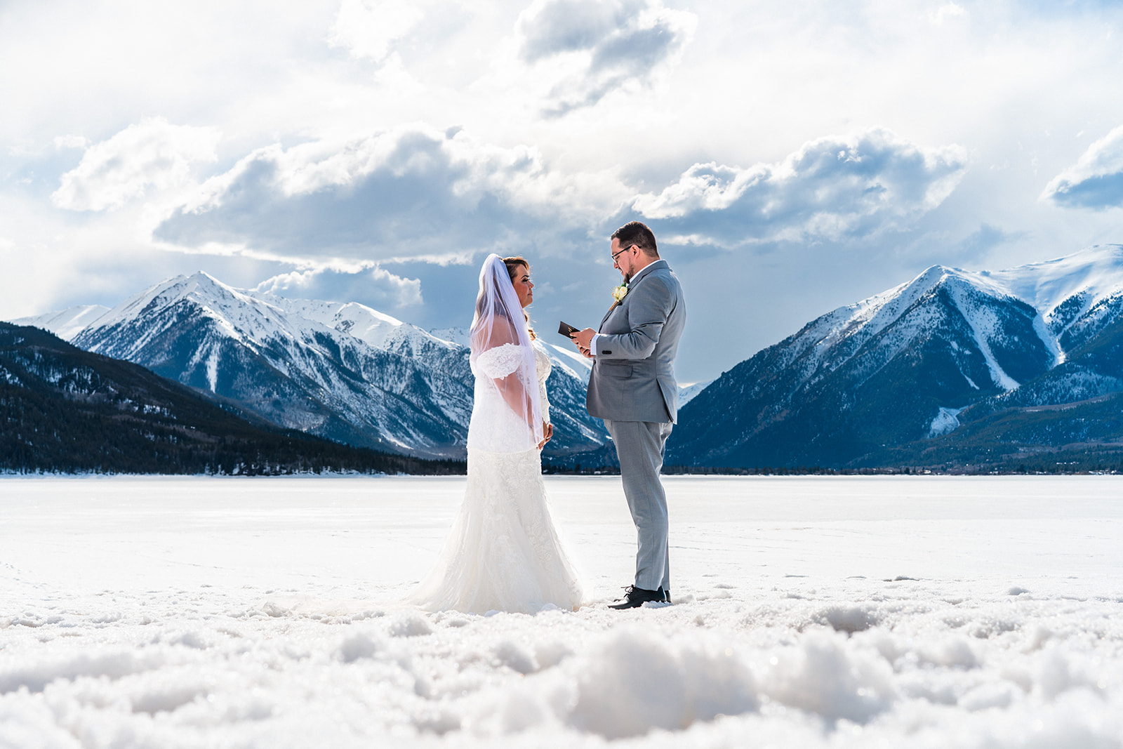 A bride and groom hold hands upon a snowy and frozen landscape with majestic mountains in the background on a sunny day beneath a cloudy sky during their Buena Vista elopement.
