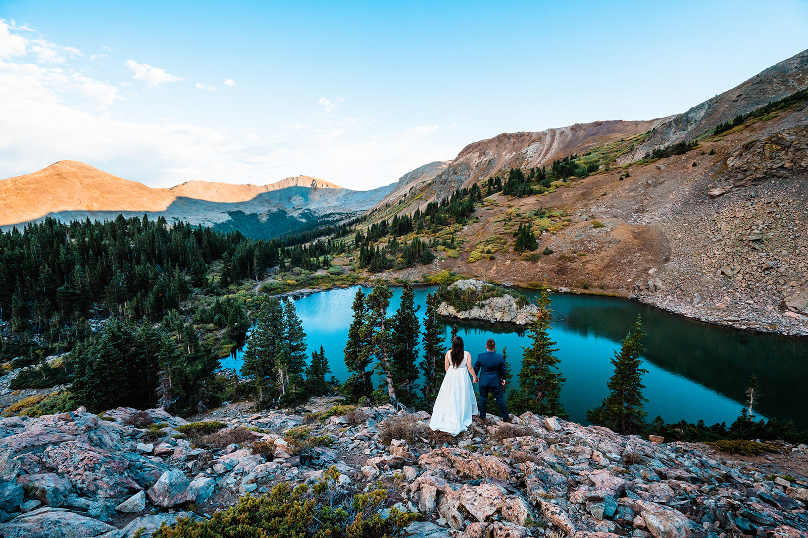 Newlyweds in formal attire holds hands, overlooking a serene blue mountain lake surrounded by lush forests and rocky peaks at sunset during their Buena Vista elopement in Colorado. 