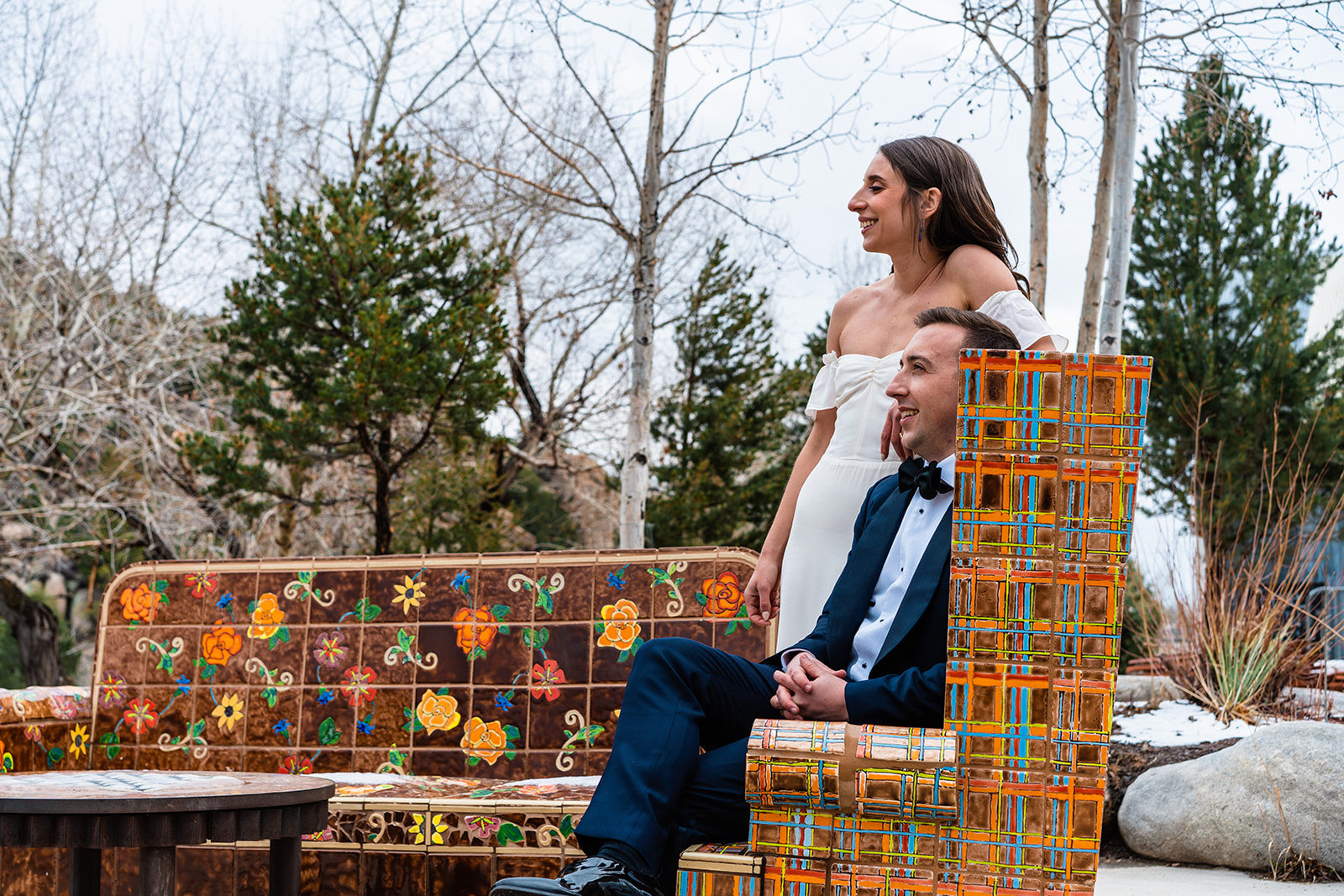 A bride and groom laugh while sitting on a colorful mosaic outdoor bench surrounded by pine trees during their Buena Vista elopement at the Surf Hotel in Colorado.