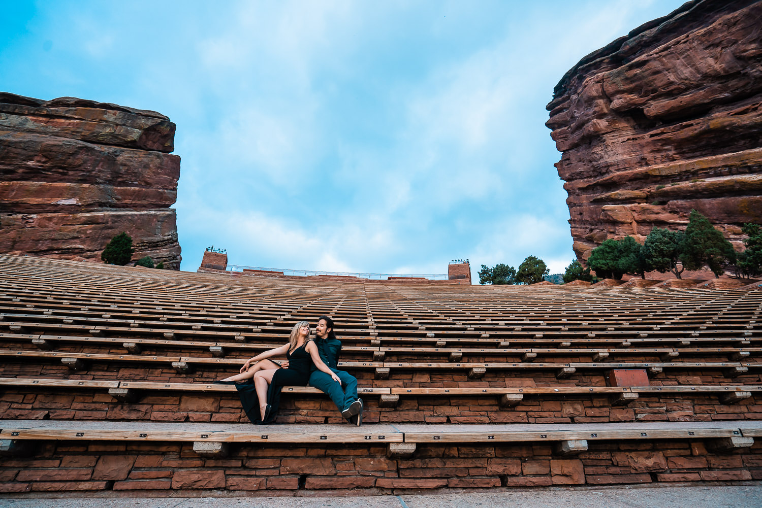 A newly engaged couple sits in the middle of an empty Red Rocks Amphitheater near Denver beneath a blue sky with puffy white clouds.