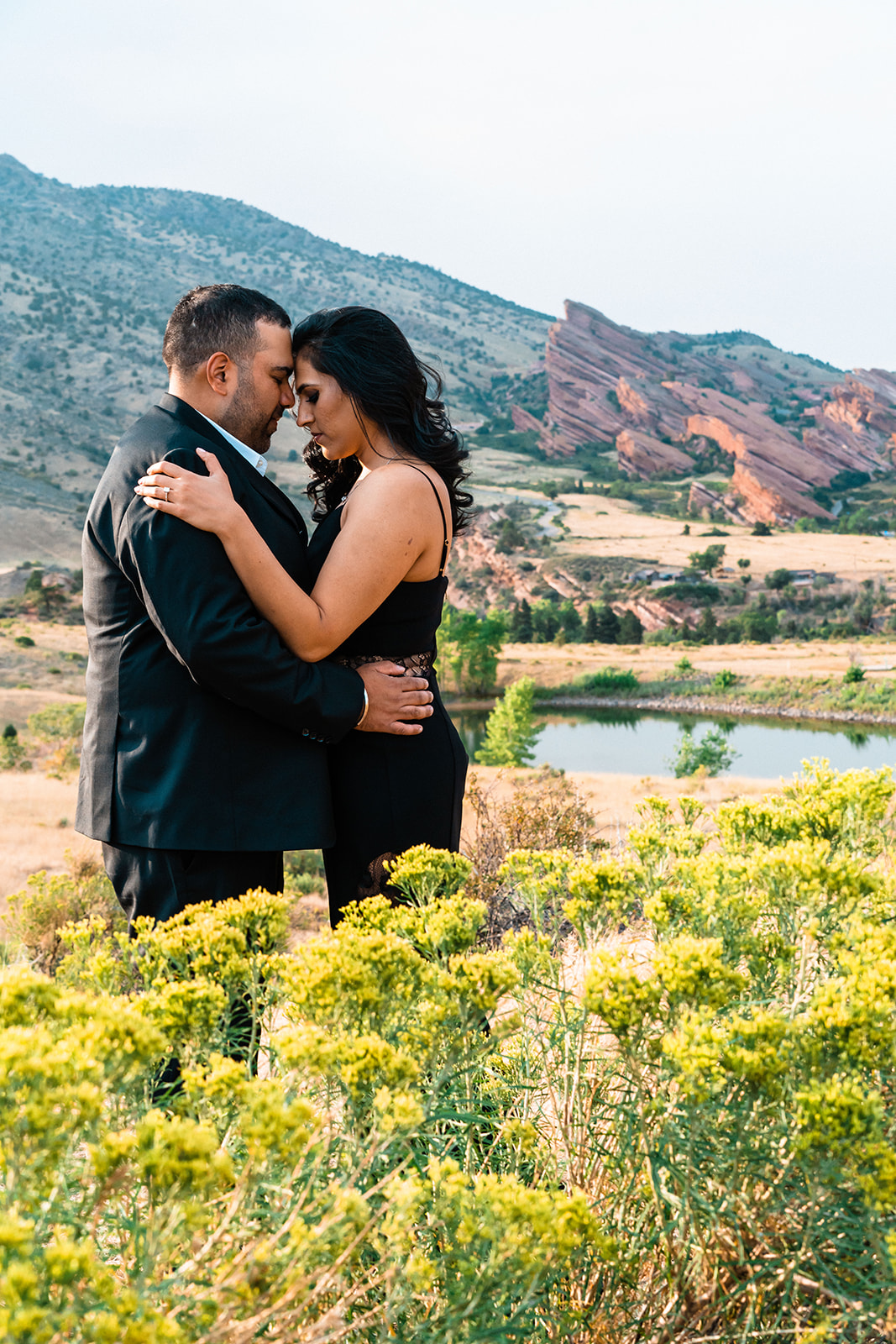 A couple embraces tenderly in a field, with a serene lake and distinctive red rock formations in the background, as featured in the Denver Elopement Guide.