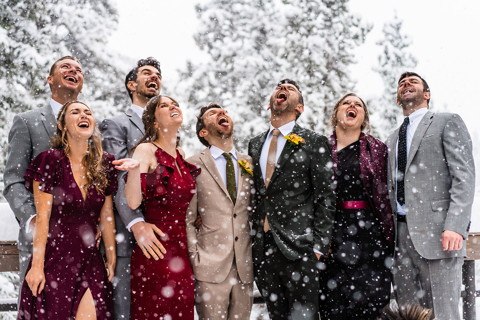 A joyful bridal party and elated grooms all in formal attire tilt their chins skyward and open their mouths to catch falling snow with snowy trees behind them as they demonstrate how to elope in Breckenridge, Colorado. 