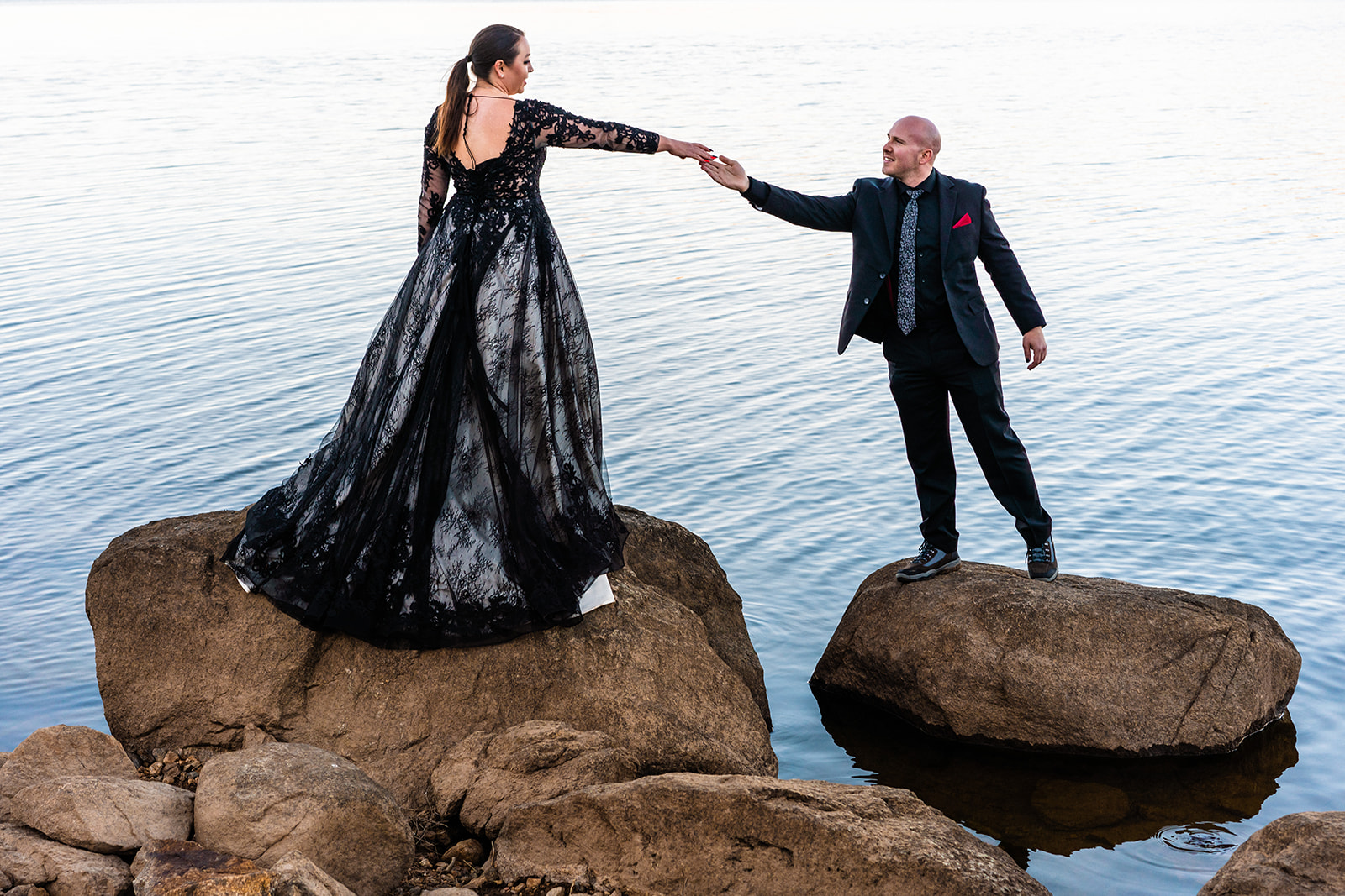 Bride and groom reaching for eachothers hands on rocks in black Halloween elopement attire