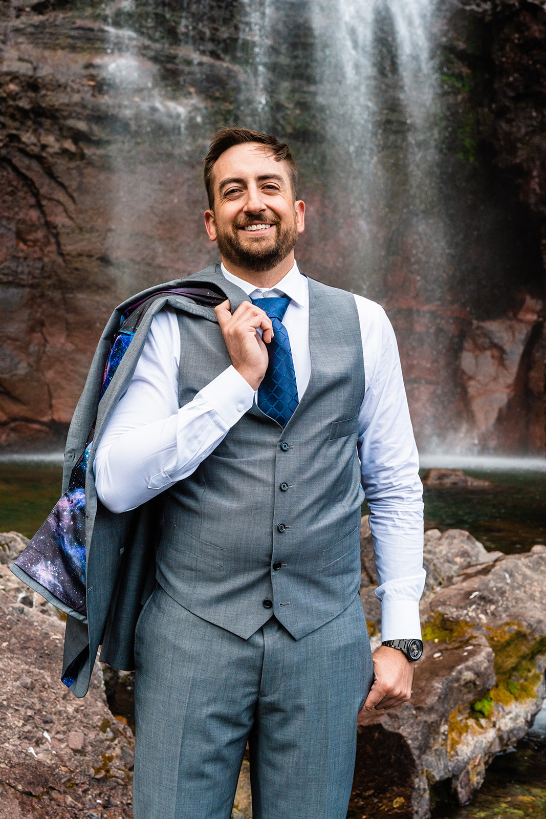Groom smiling posing in front of Bridal Veil falls in a grey suite