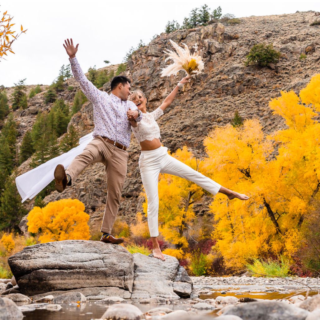 A couple jumping with their arms and legs in the air in front of a Colorado mountain forest