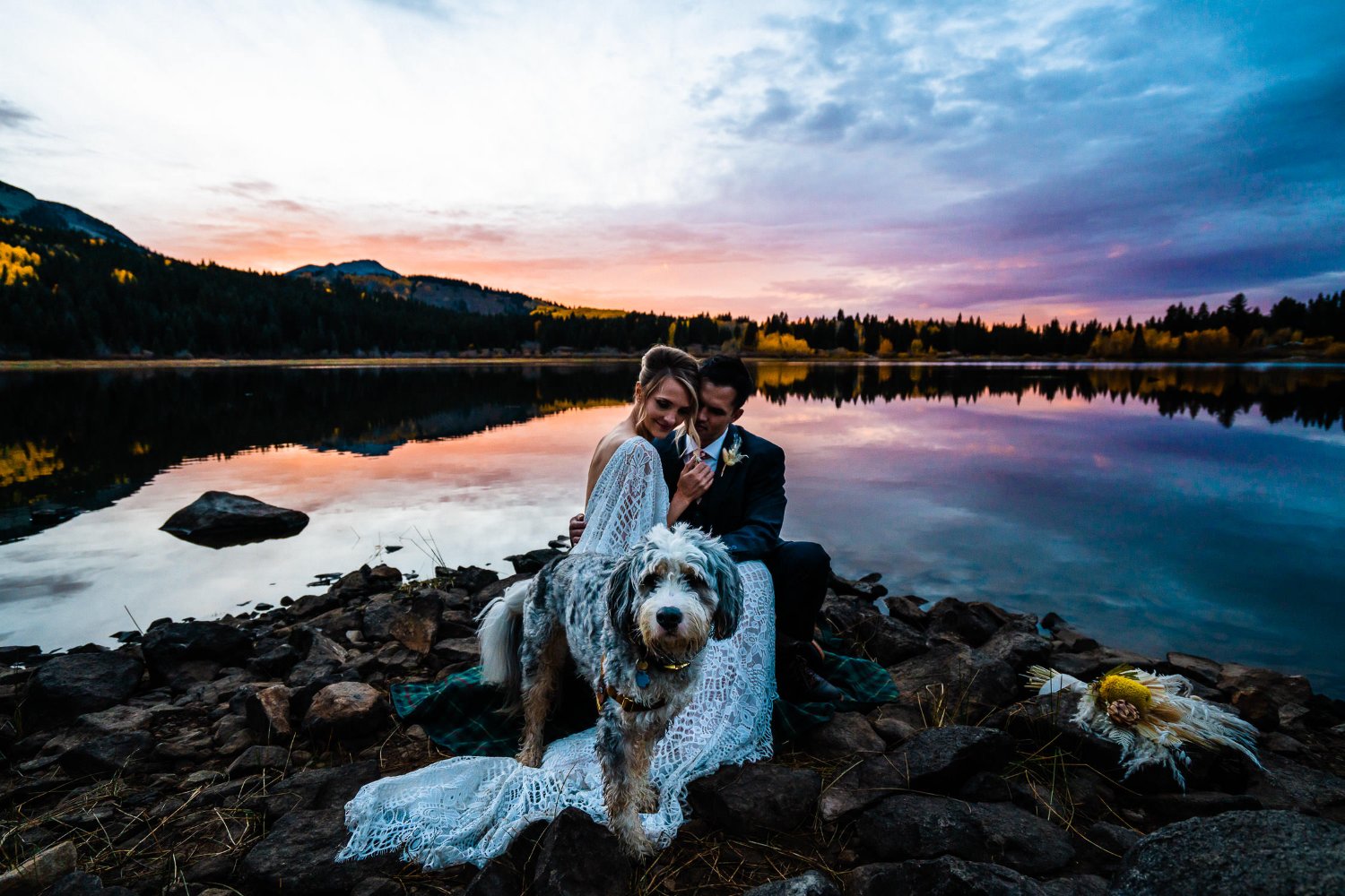 Elopement photos of a bride and groom sitting on rocks by a lake with their dog.