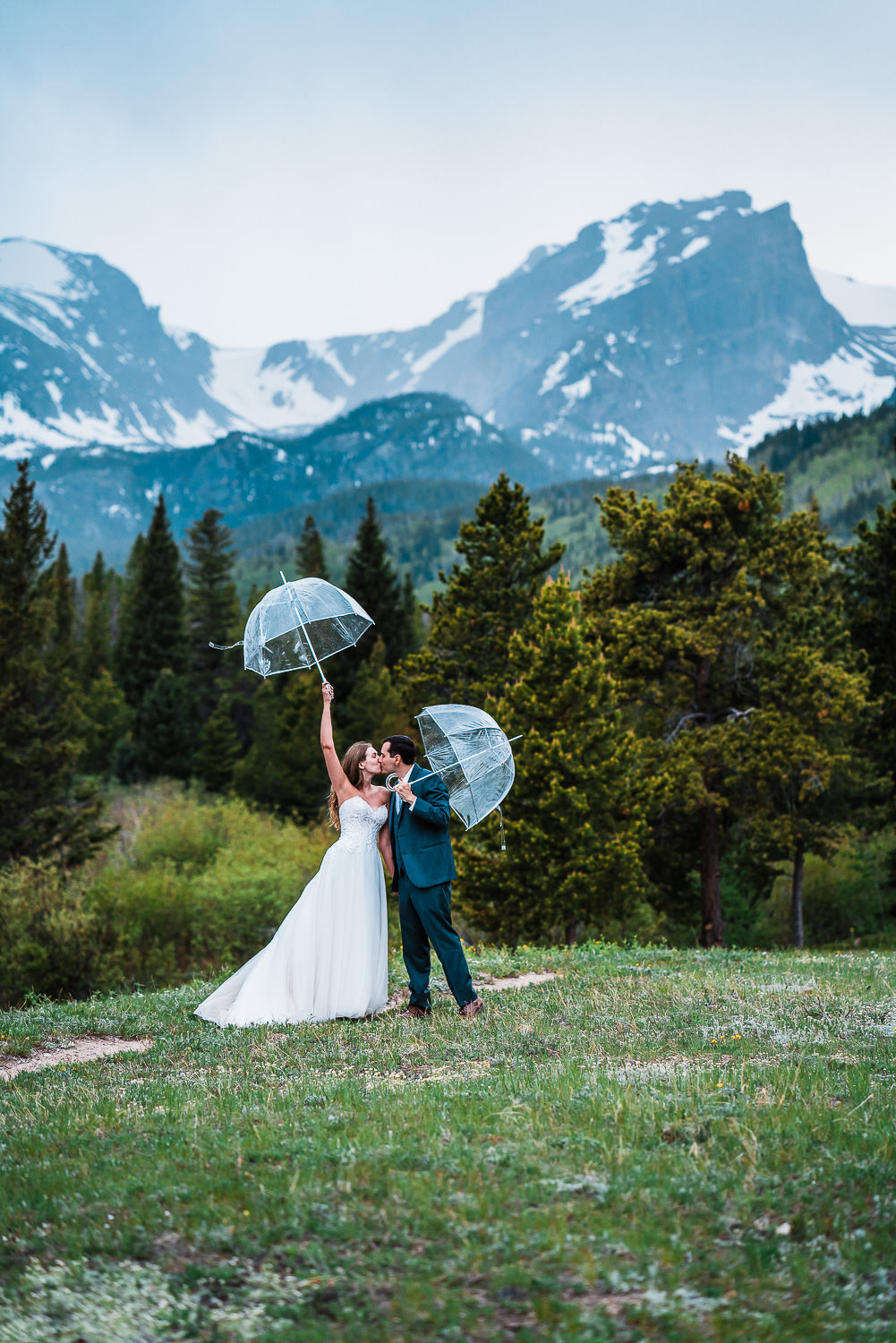 a bride and groom kiss holding umbrellas with mountains and trees behind them