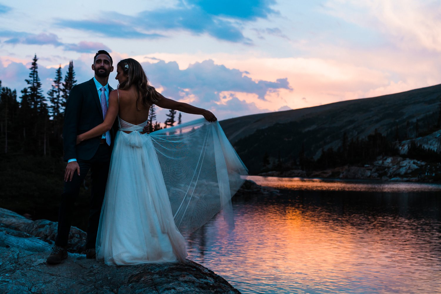 An elopement couple standing on rocks near a lake at sunset, capturing stunning photos.