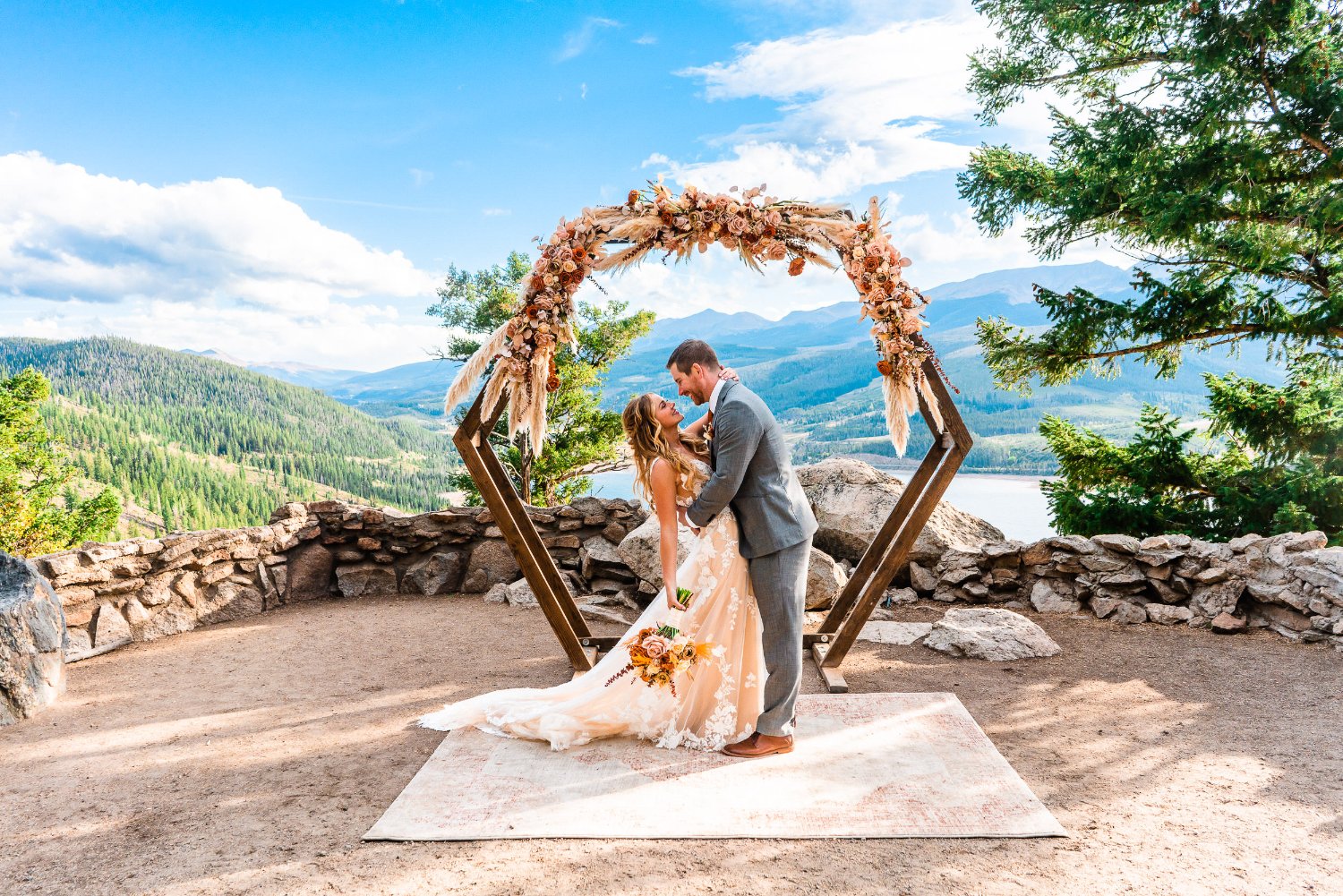 An intimate elopement photoshoot captures a bride and groom sharing a romantic kiss under a breathtaking arch set amidst majestic mountains.