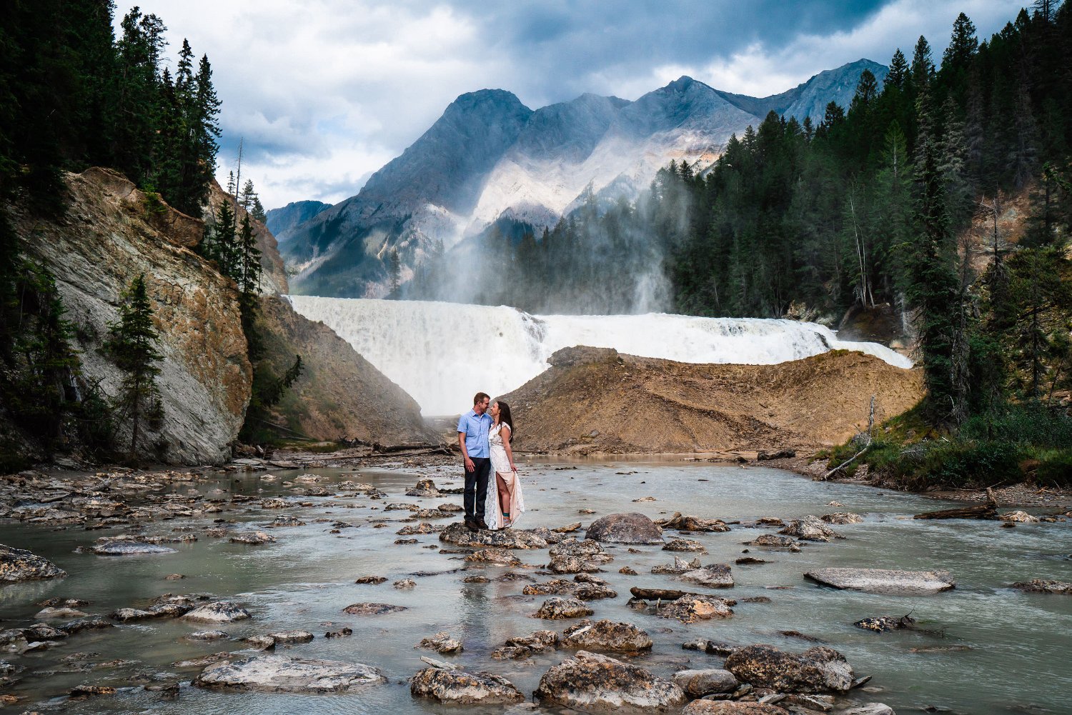An adventurous couple captures elopement photos in front of a breathtaking waterfall, with majestic mountains as the backdrop.