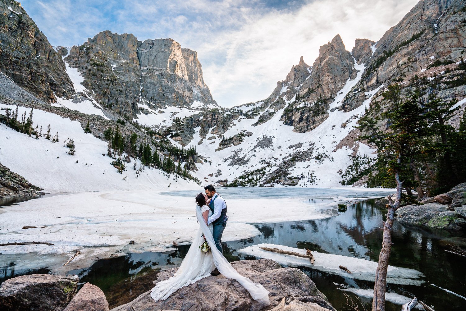 Stunning elopement photos of a bride and groom standing on rocks near a picturesque lake in the mountains.