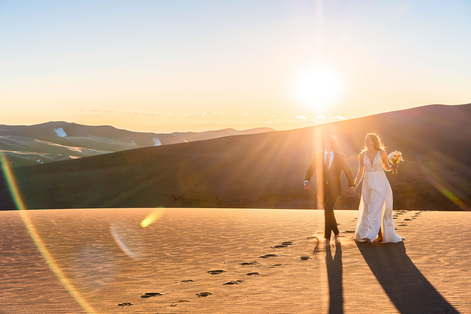 A bride and groom walking through the sand dunes at sunset.