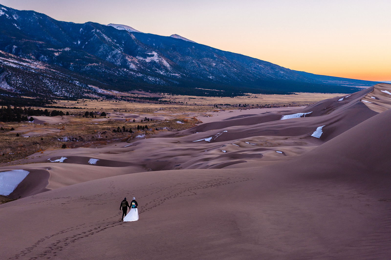 Bride and groom walking down a sandy hill at the Great Sand Dunes with mountains in the background.