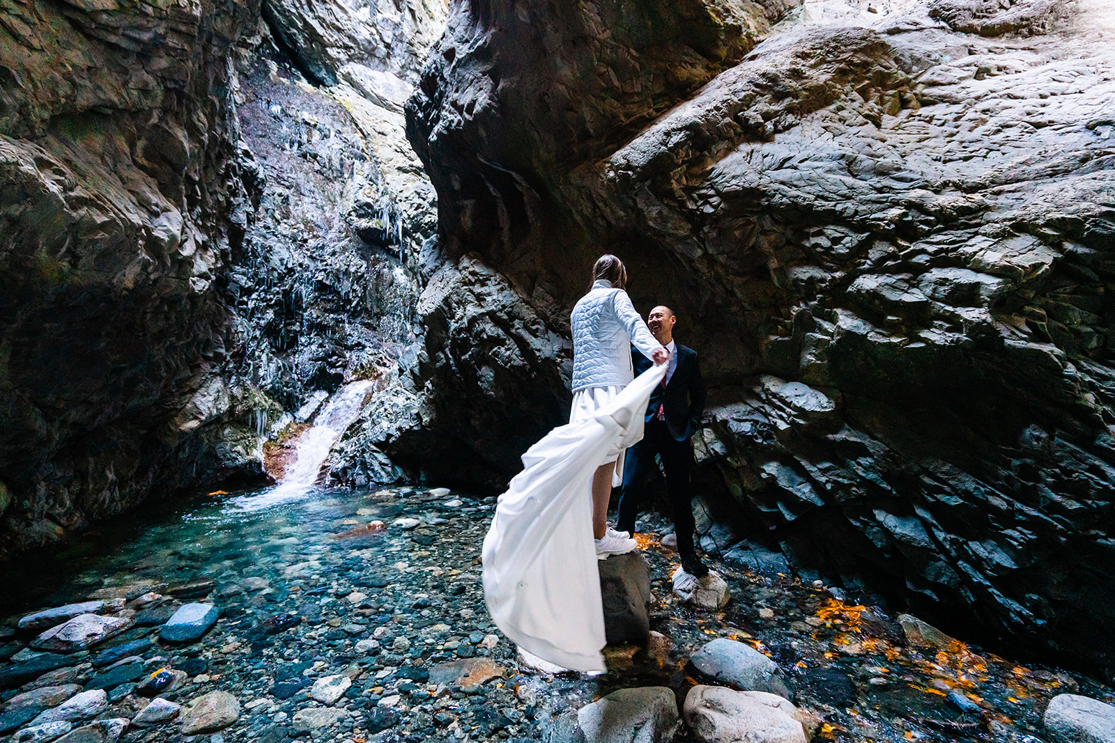A bride and groom standing next to a waterfall in a canyon.