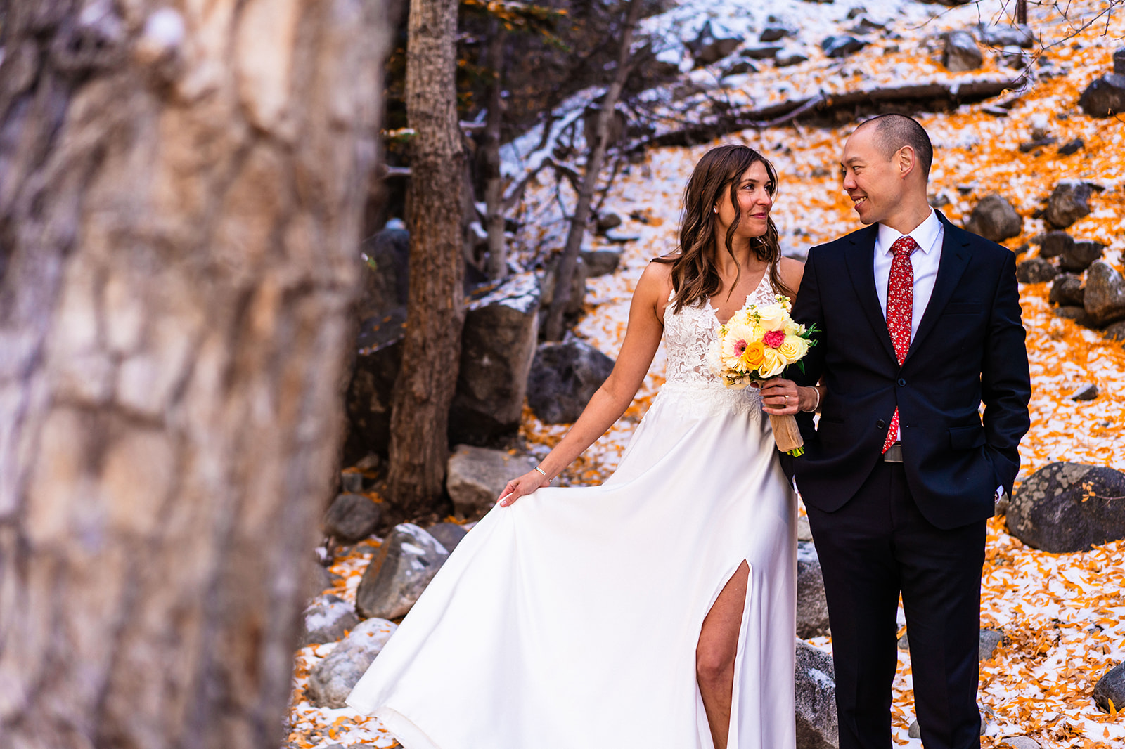 A bride and groom standing in front of a tree in the snow smiling at one another.