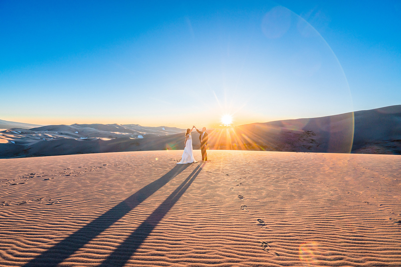 A bride and groom standing on a sand dune at sunset.