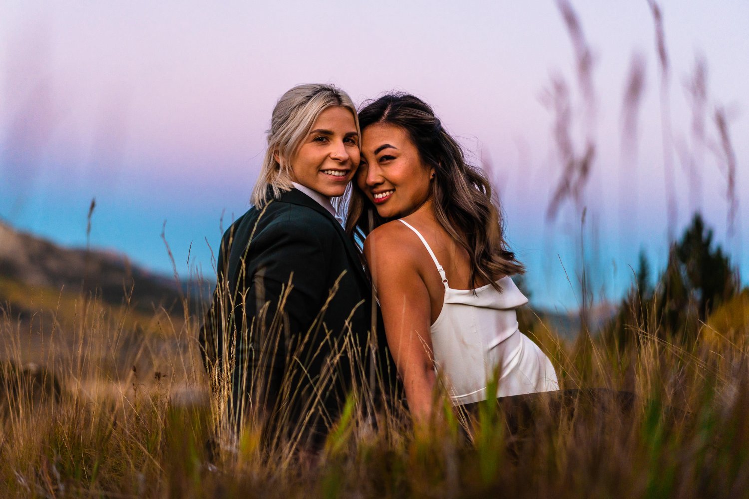 Two women capturing intimate elopement photos in tall grass at sunset.