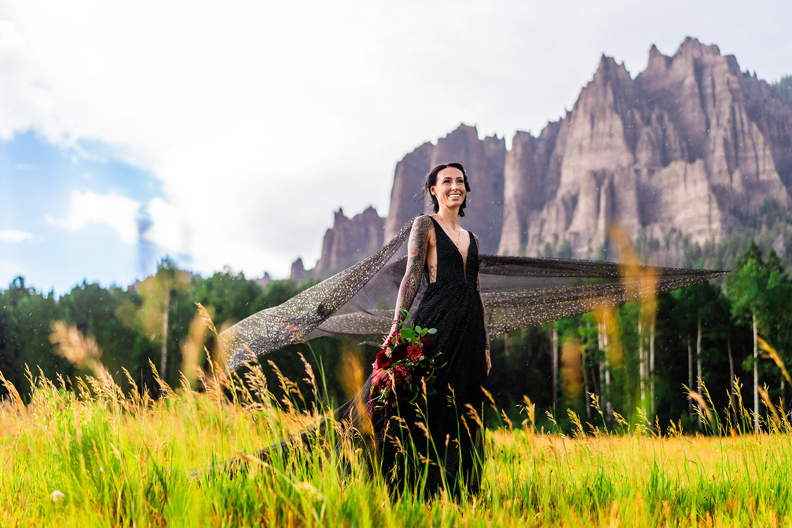 A bride in a black elopement dress standing in a field with mountains in the background.