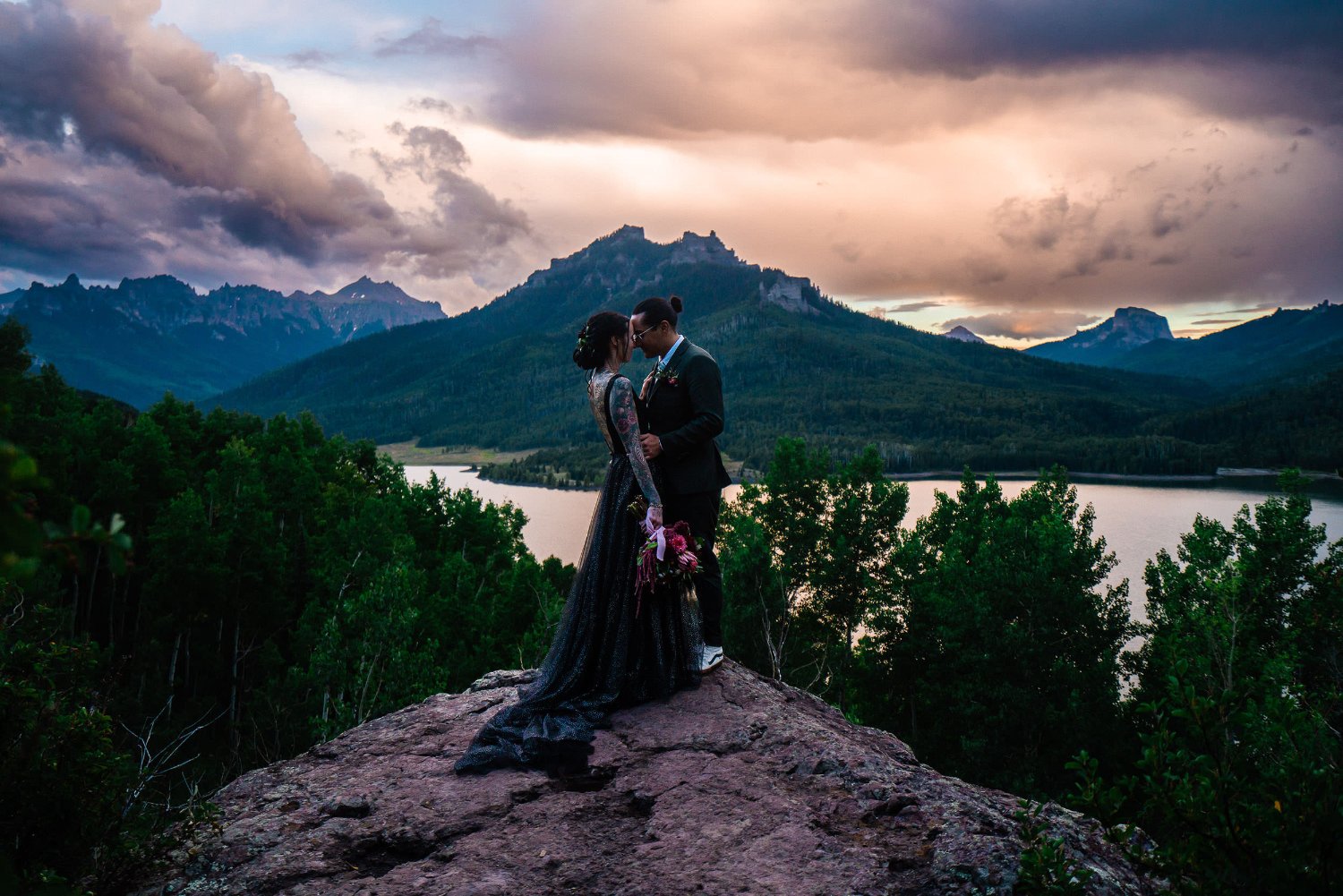 A blissful elopement photoshoot featuring a loving man and woman sharing a passionate kiss on a majestic rock amidst breathtaking mountains and tranquil water.