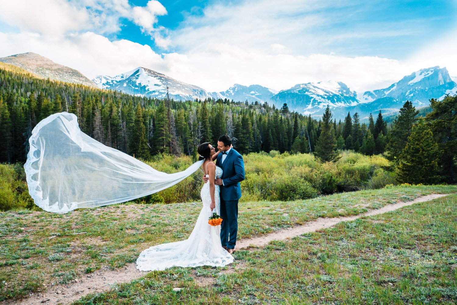A bride and groom in their elopement photos share a romantic kiss in front of the majestic mountains, as their veil gracefully blows in the wind.