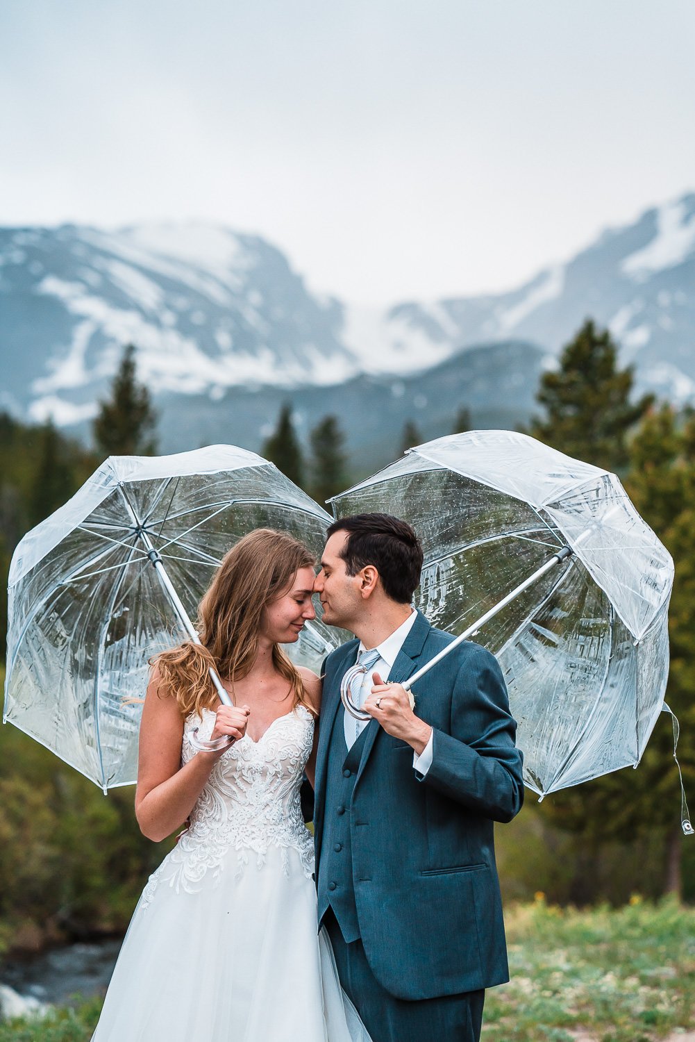 Elopement photos of a bride and groom sharing a romantic kiss under umbrellas in front of a majestic mountain.