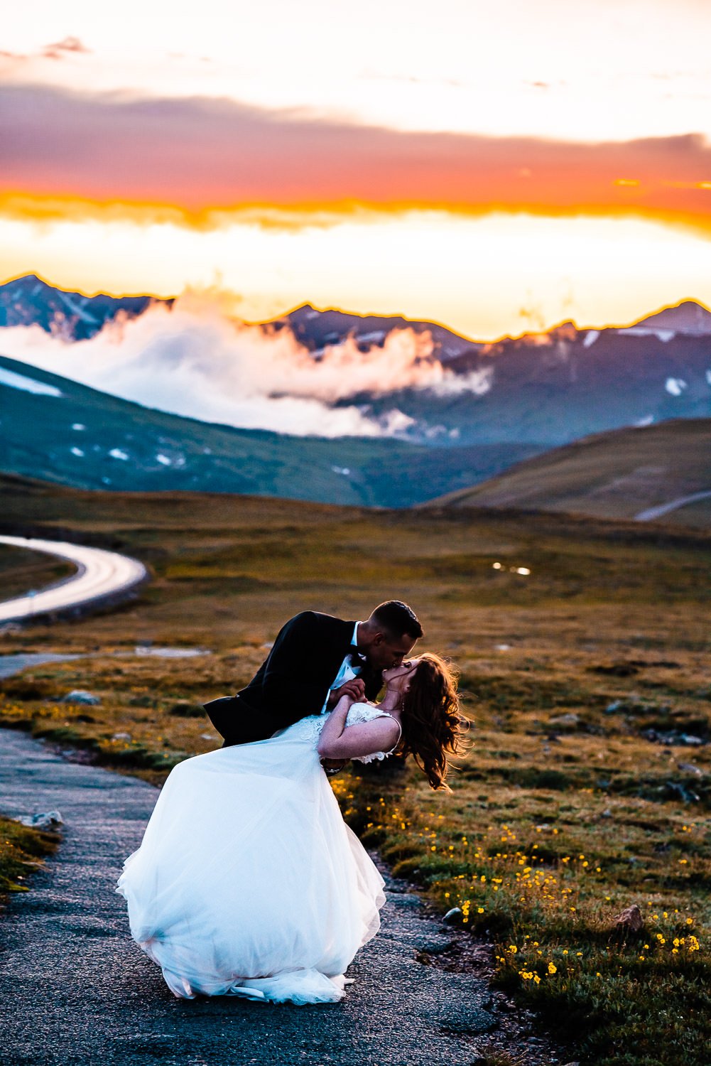 A couple embraces in a breathtaking elopement photoshoot, sharing a passionate kiss on a scenic road with majestic mountains as their backdrop.