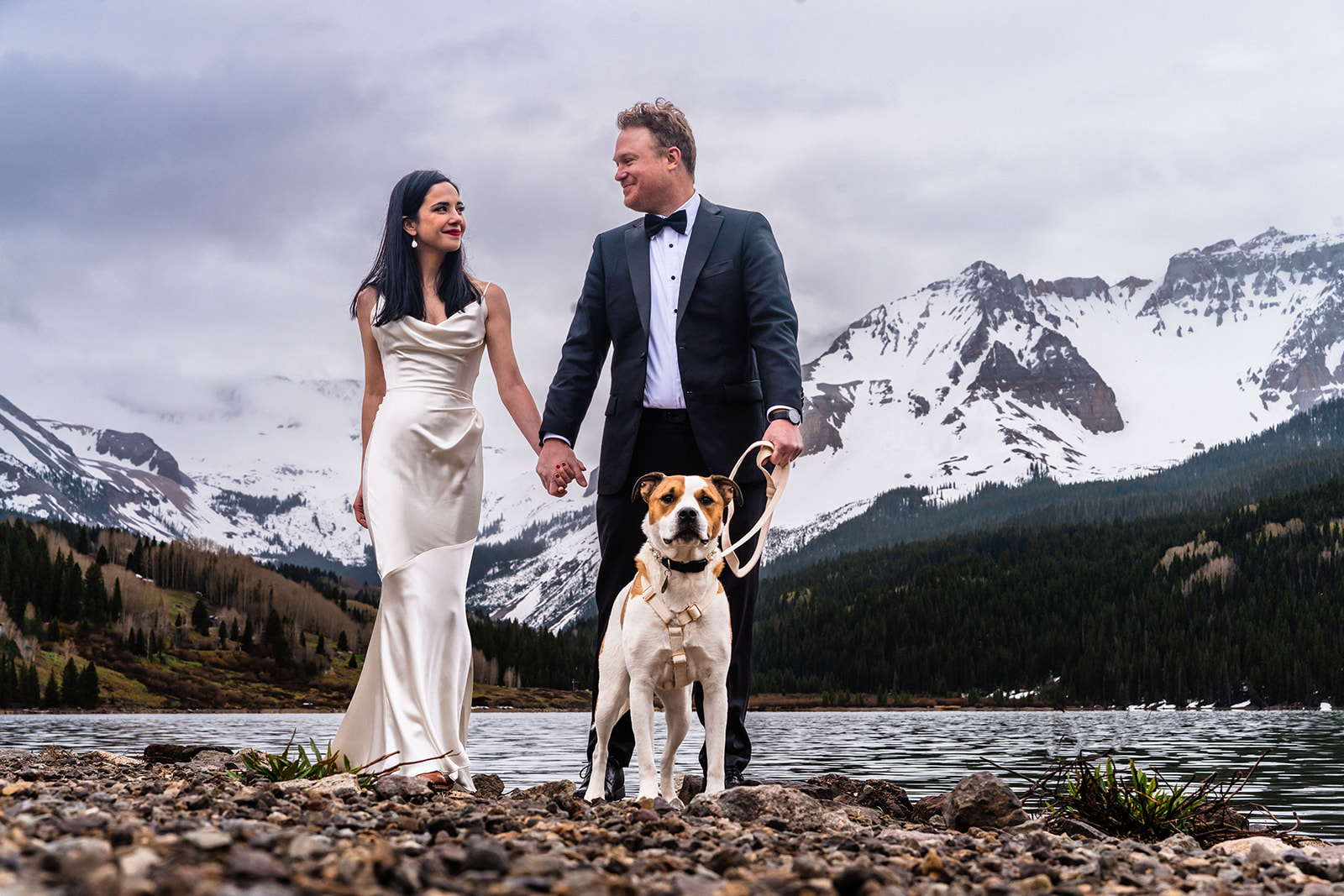 A bride and groom standing next to a lake with mountains in the background.