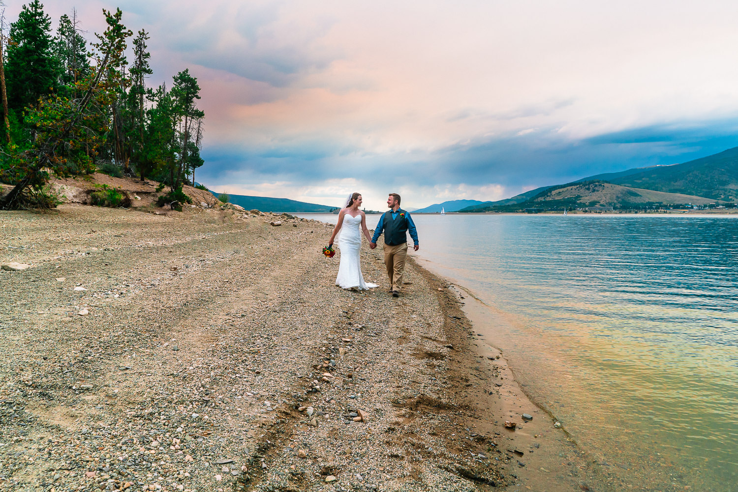 A bride and groom hold hands and walk along the shoreline of Lake Dillon in Breckenridge, Colorado during their Windy Point Campground Wedding.