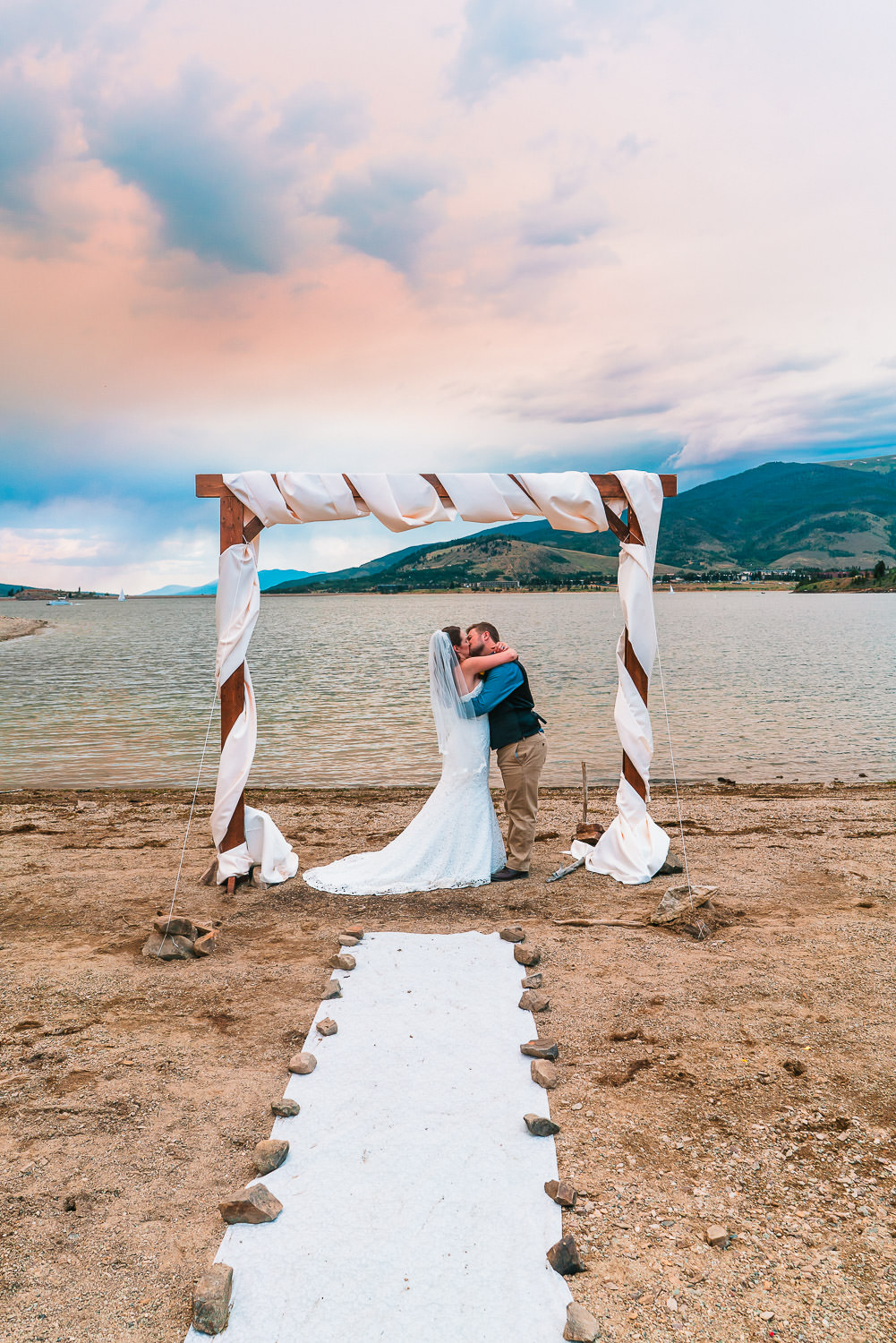 A bride and groom kiss beneath their altar on the shoreline of Lake Dillon in Breckenridge, Colorado during their Windy Point Campground Wedding.
