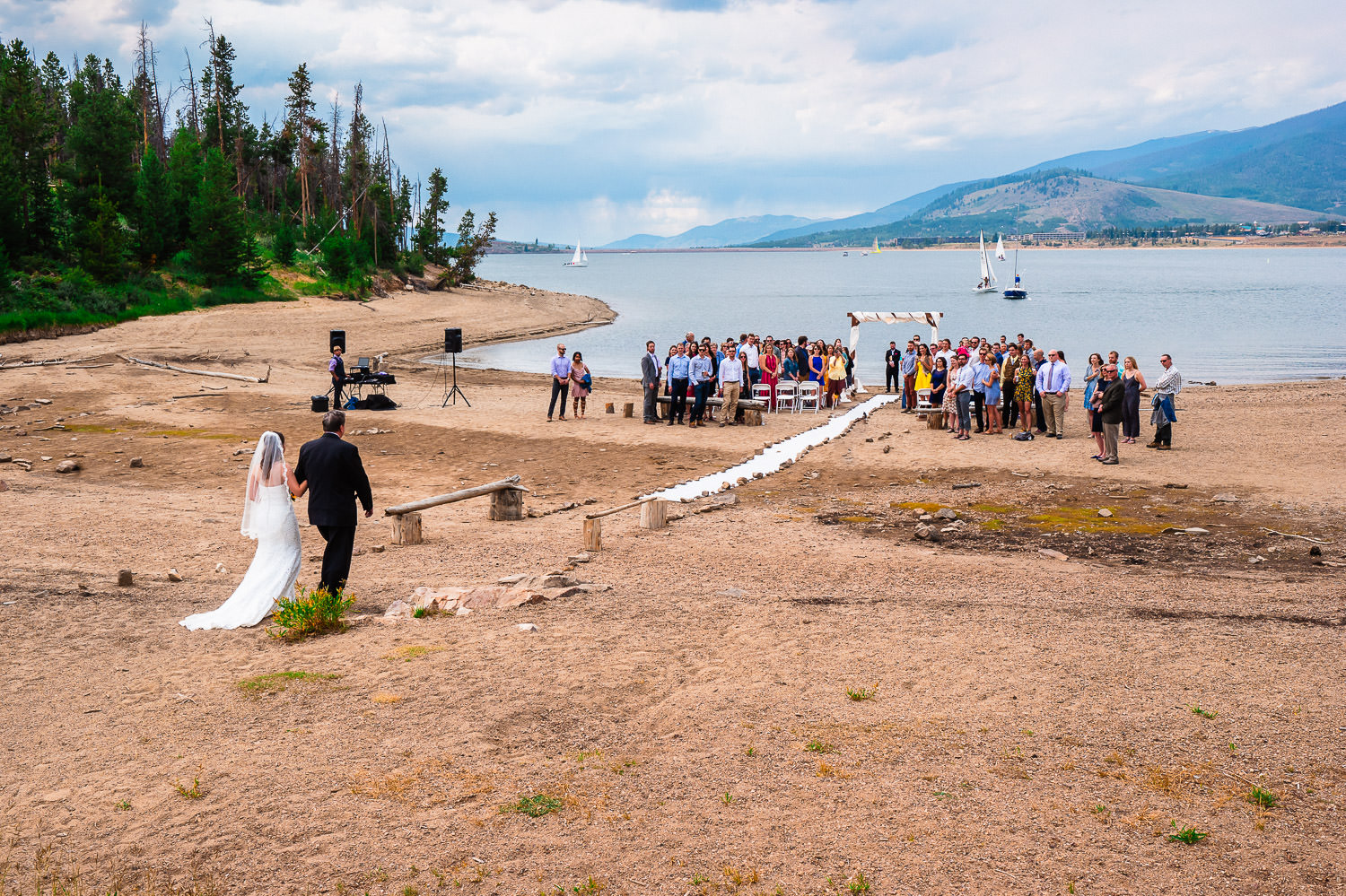 A bride and groom walk to their ceremony altar during their Windy Point Campground Wedding with sailboats sailing upon Lake Dillon in the distance