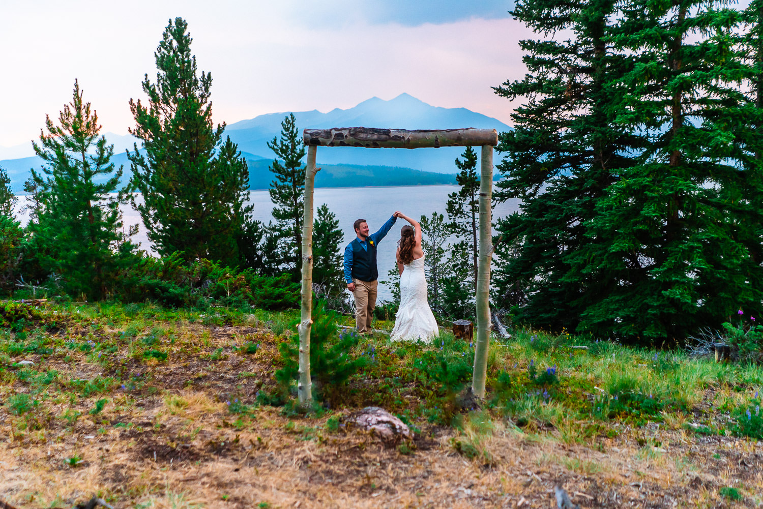 A couple in wedding attire holding hands and dancing under a rustic wooden frame with the mountainous landscape of Lake Dillon in the background, celebrating during their Windy Point Campground wedding.