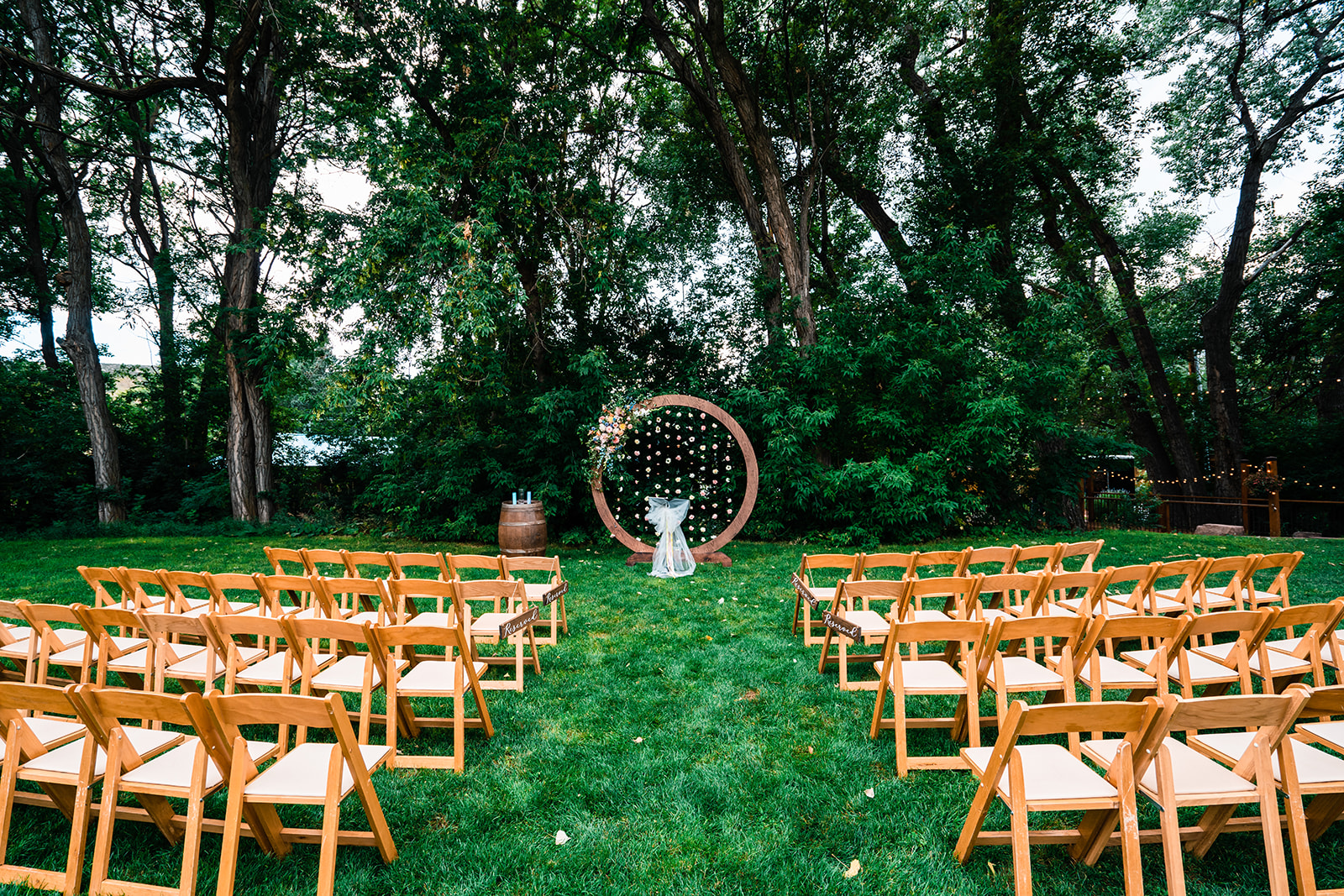 Outdoor wedding ceremony site setup with rows of wooden chairs facing a decorative circular arch in a grassy area surrounded by trees.