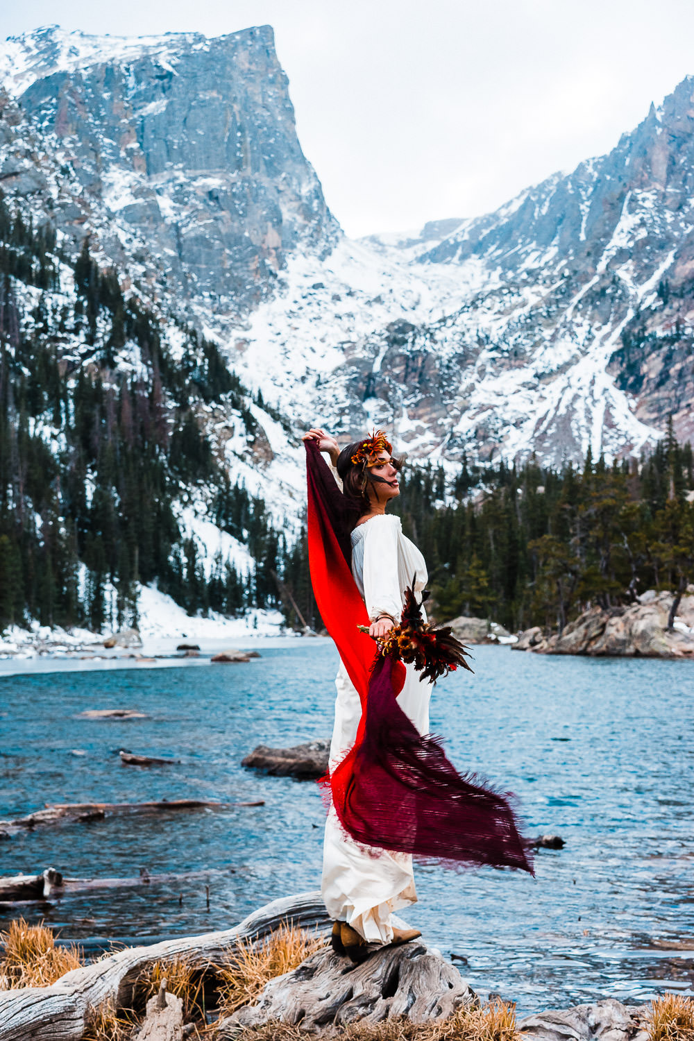 A woman in a white mountain elopement dress with a red scarf is standing on a rock next to a lake during her Colorado elopement.