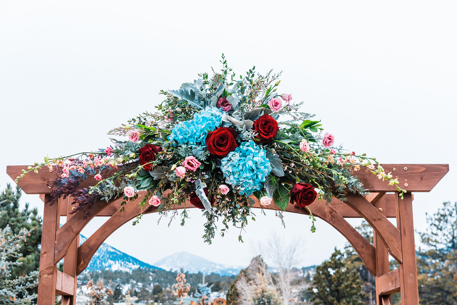 Bright blue, red, and green floral arrangement atop a wood square wedding arch with a snowy mountain landscape in the background.