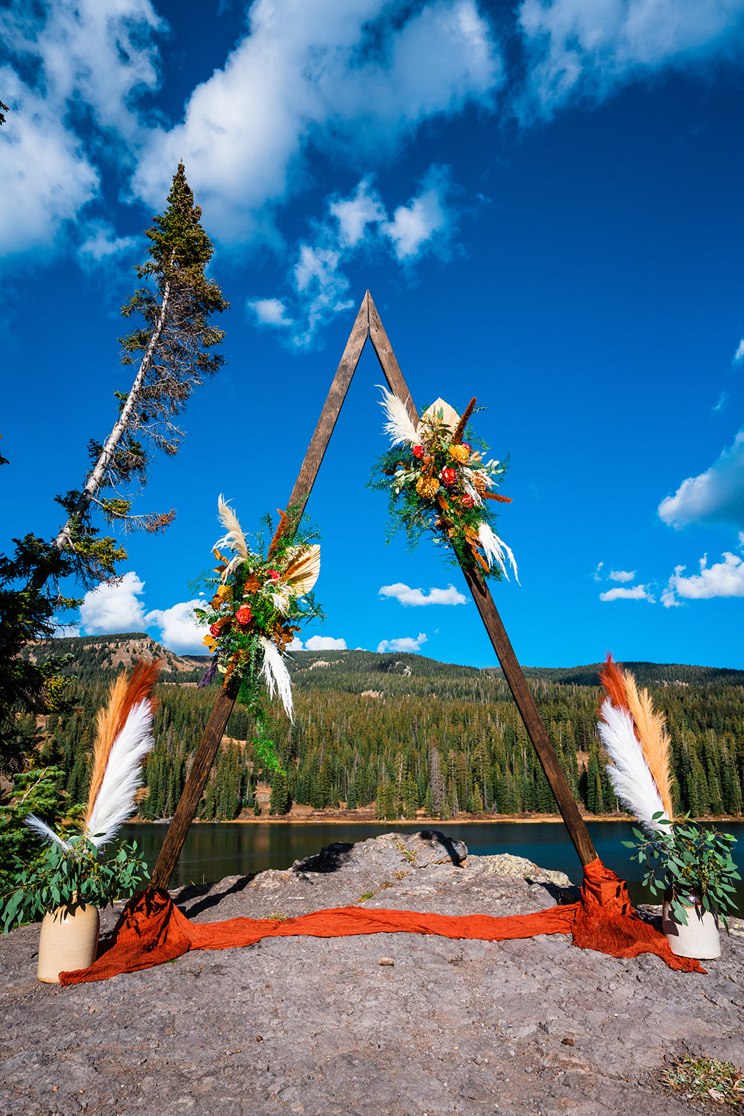 Triangle wedding arch for an outdoor elopement ceremony adorned with multiple bright and colorful floral arrangements, set against a backdrop of a deep blue sky, a few puffy white clouds, and a serene mountain lake in Colorado