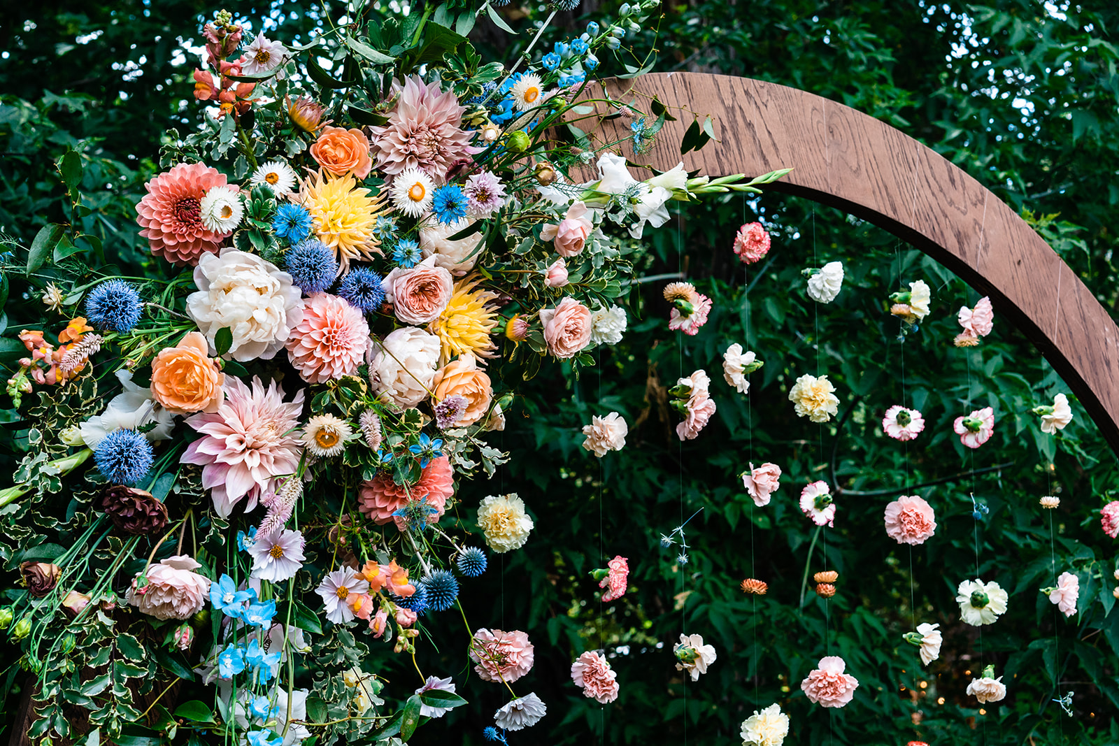 A colorful floral arrangement on a circular wooden arch with flowers suspended in mid-air against a backdrop of green foliage that sparks incredible wedding ceremony arch ideas.