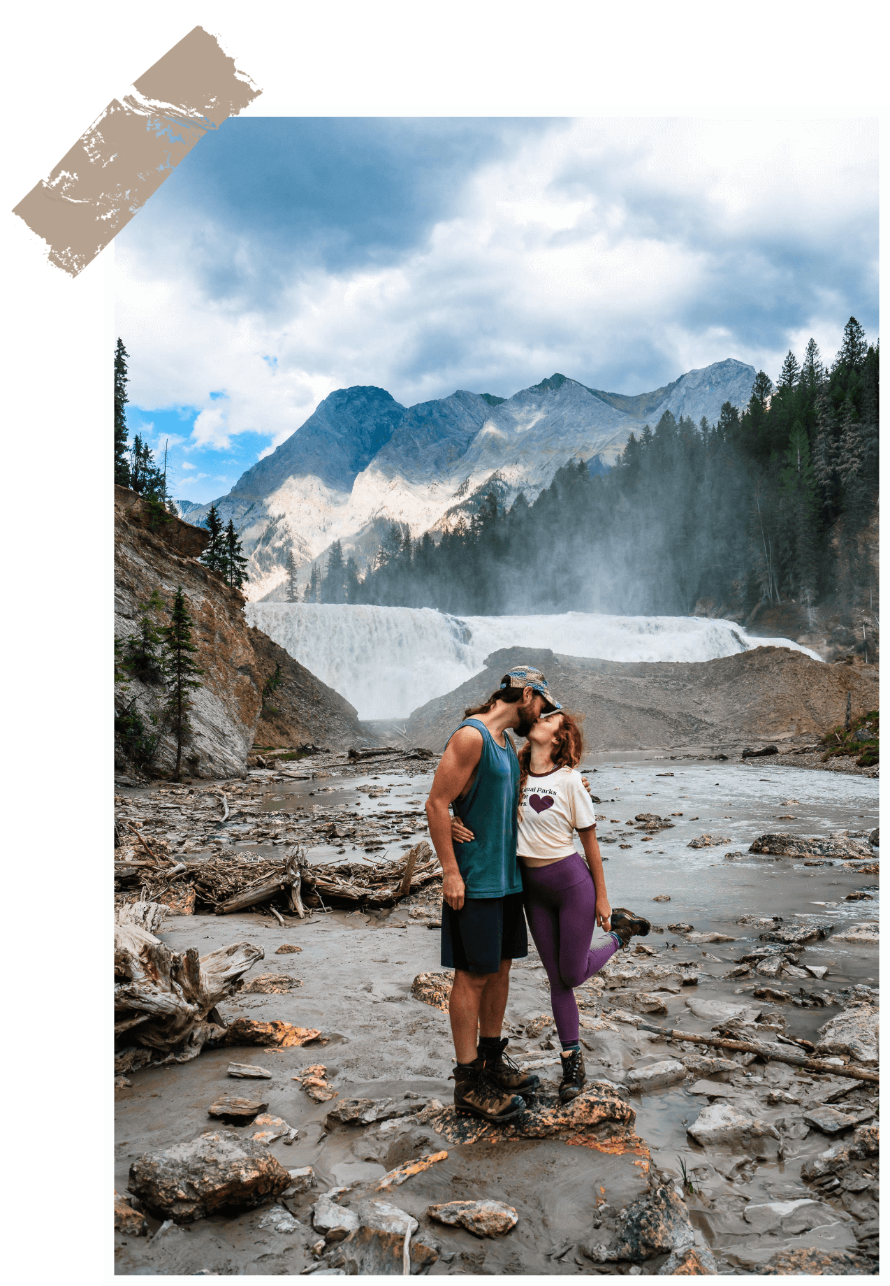 Meg and Kevin sharing a kiss on a rocky riverbed with a waterfall and mountains in the background.